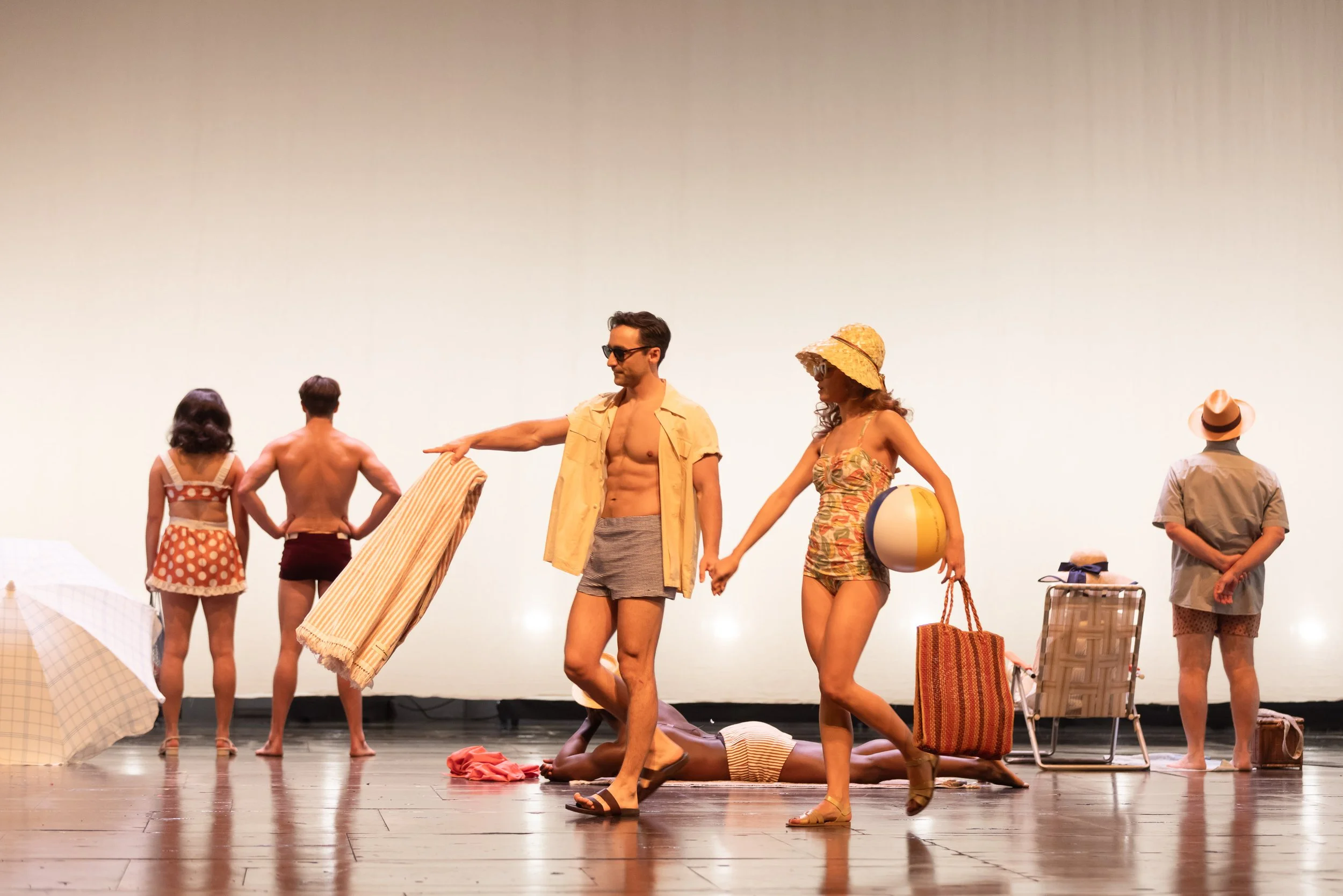 The image shows a group of people on a stage, some dressed in vintage swimwear, holding hands, and engaging in a performance or rehearsal. There are beach accessories such as umbrellas, a beach ball, and chairs scattered around the scene.