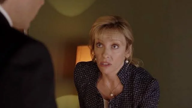 A woman with short blonde hair looking at a man, with a concerned or serious expression, in a dimly lit room.