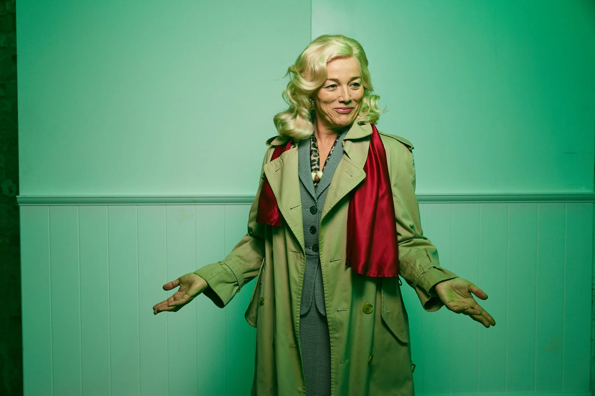 An older woman with blonde, wavy hair standing against a light green wall, wearing a tan trench coat with a red scarf and a patterned blouse. She is smiling softly with her eyes slightly closed, and her hands are open with palms facing upward.