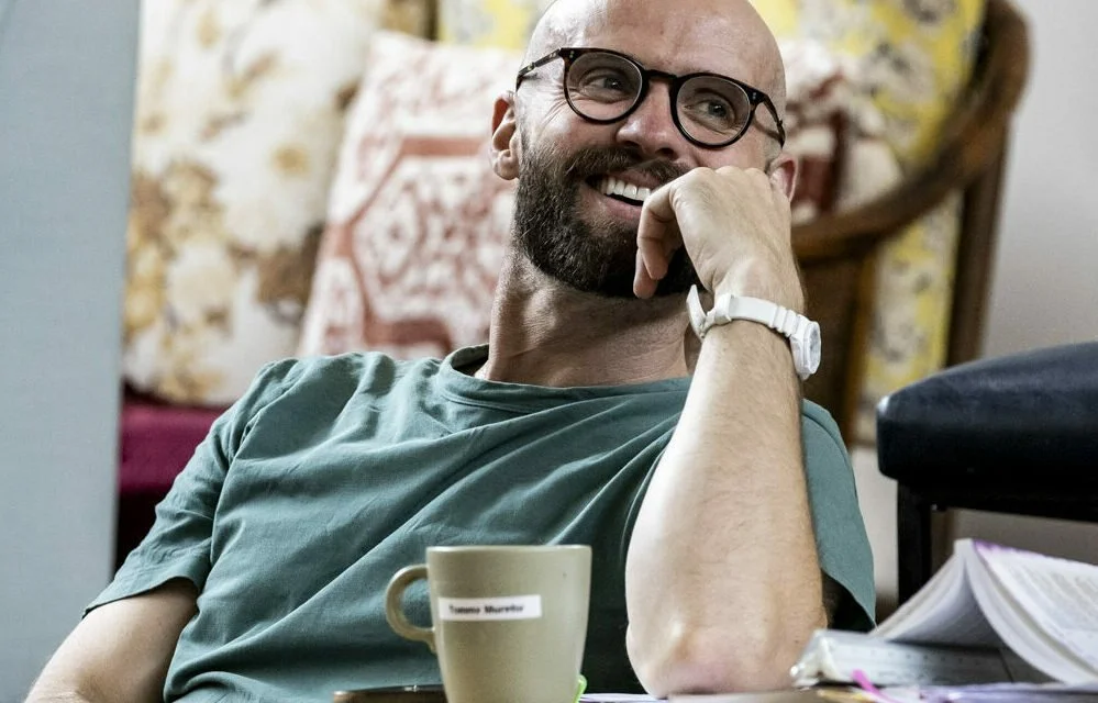 Man with glasses and a beard smiling, sitting at a table with a cup in front of him.