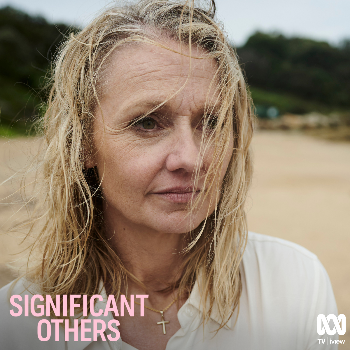 Close-up of a woman with blonde, wavy hair standing on a beach, with wind blowing her hair, wearing a white shirt and a gold cross necklace, with a blurred background of the beach and greenery.
