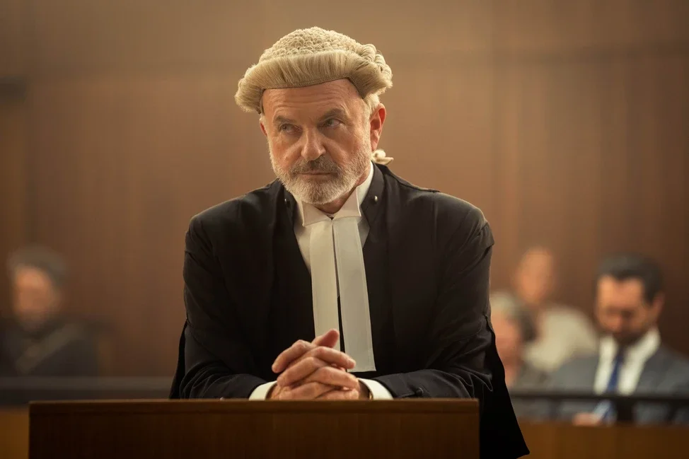A judge wearing a white wig and black robes sitting in a courtroom with his hands clasped in front of him, with people seated in the background.