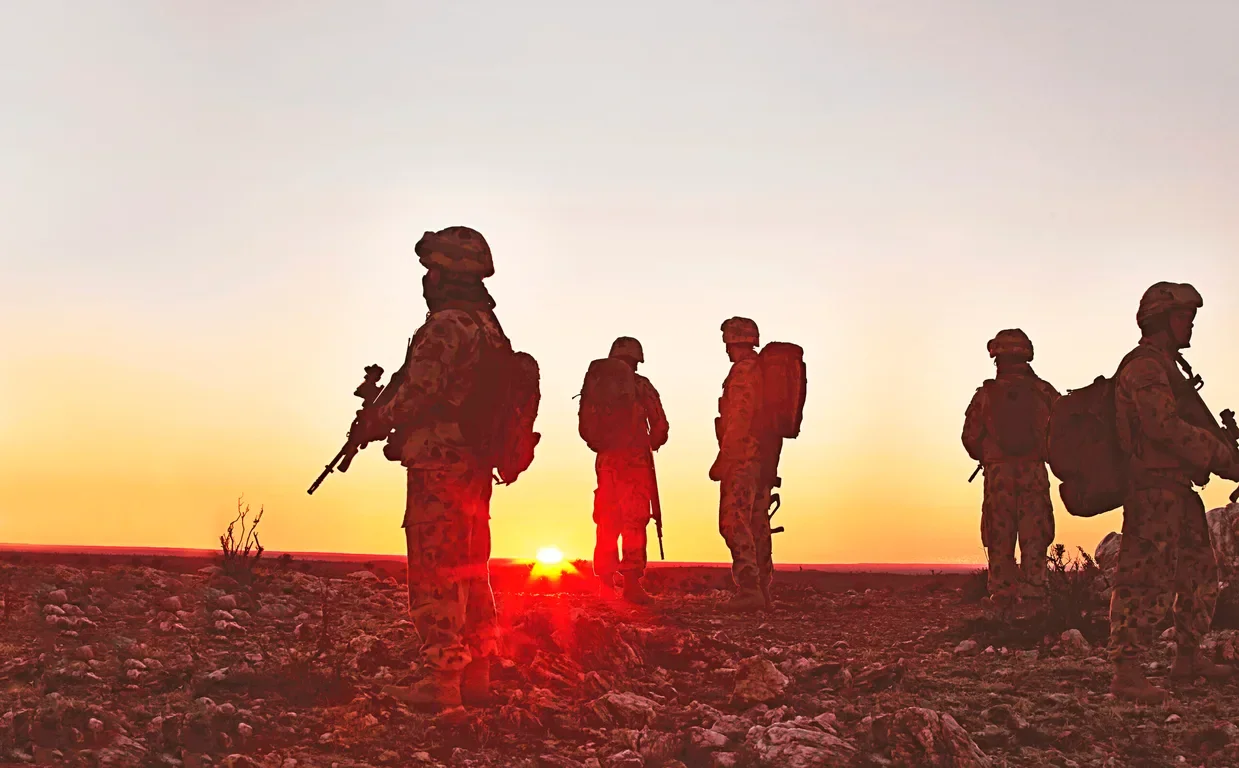 Silhouettes of six soldiers standing on rocky terrain at sunset, some holding rifles, some with backpacks.