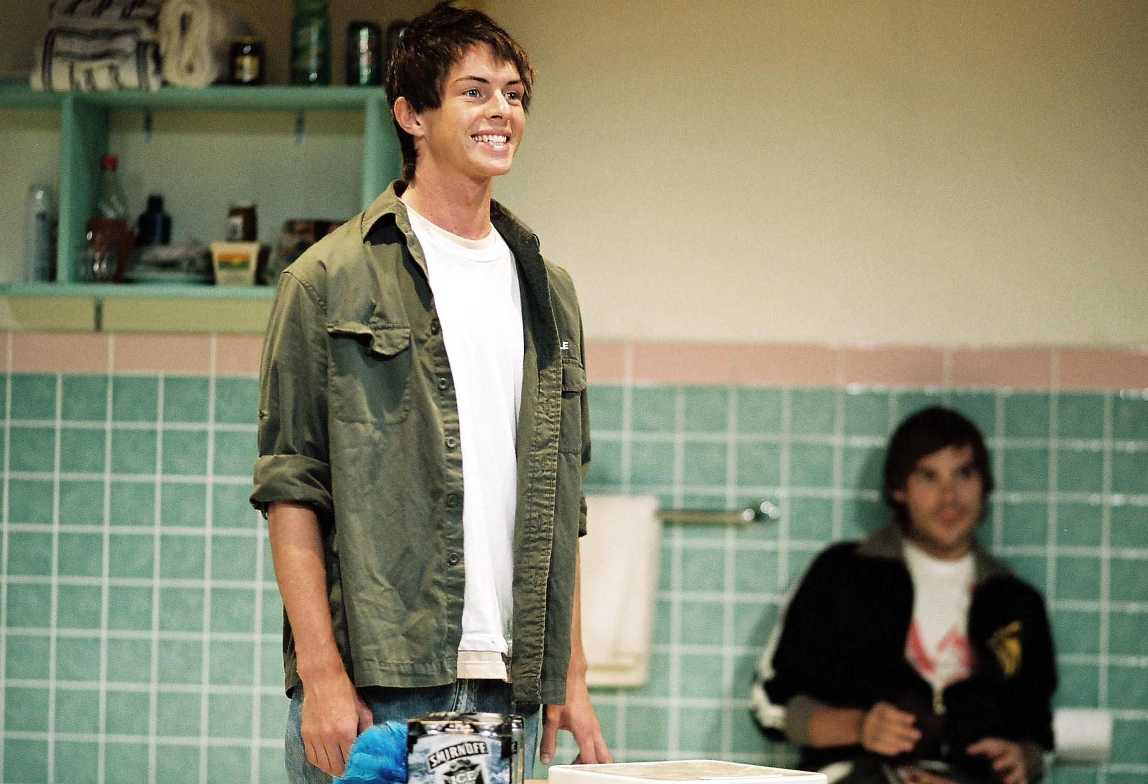 A young man with brown hair, wearing a green jacket and white T-shirt, standing in a room with green tiles on the wall, smiling and holding a box of snacks, with a woman sitting in the background.
