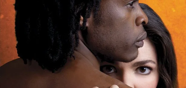 A close-up of a man and woman embracing with the woman's face partially hidden behind the man's shoulder, and both looking serious against an orange background.