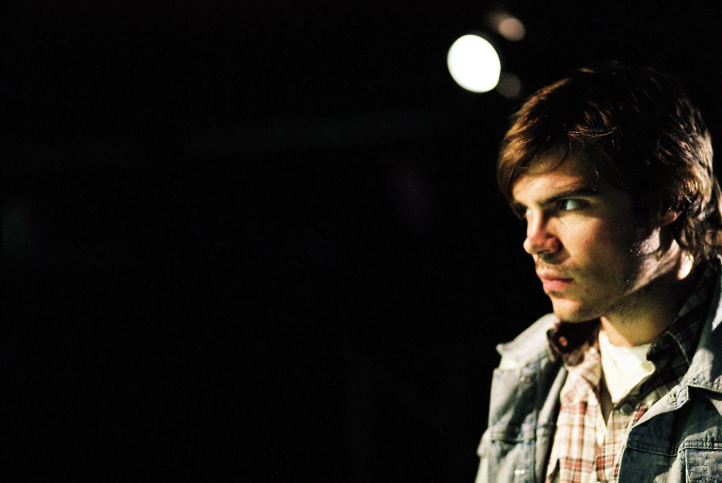 Close-up of a young man's face with curly hair, wearing a checkered shirt and denim jacket, looking to the side against a dark background with a light source behind.