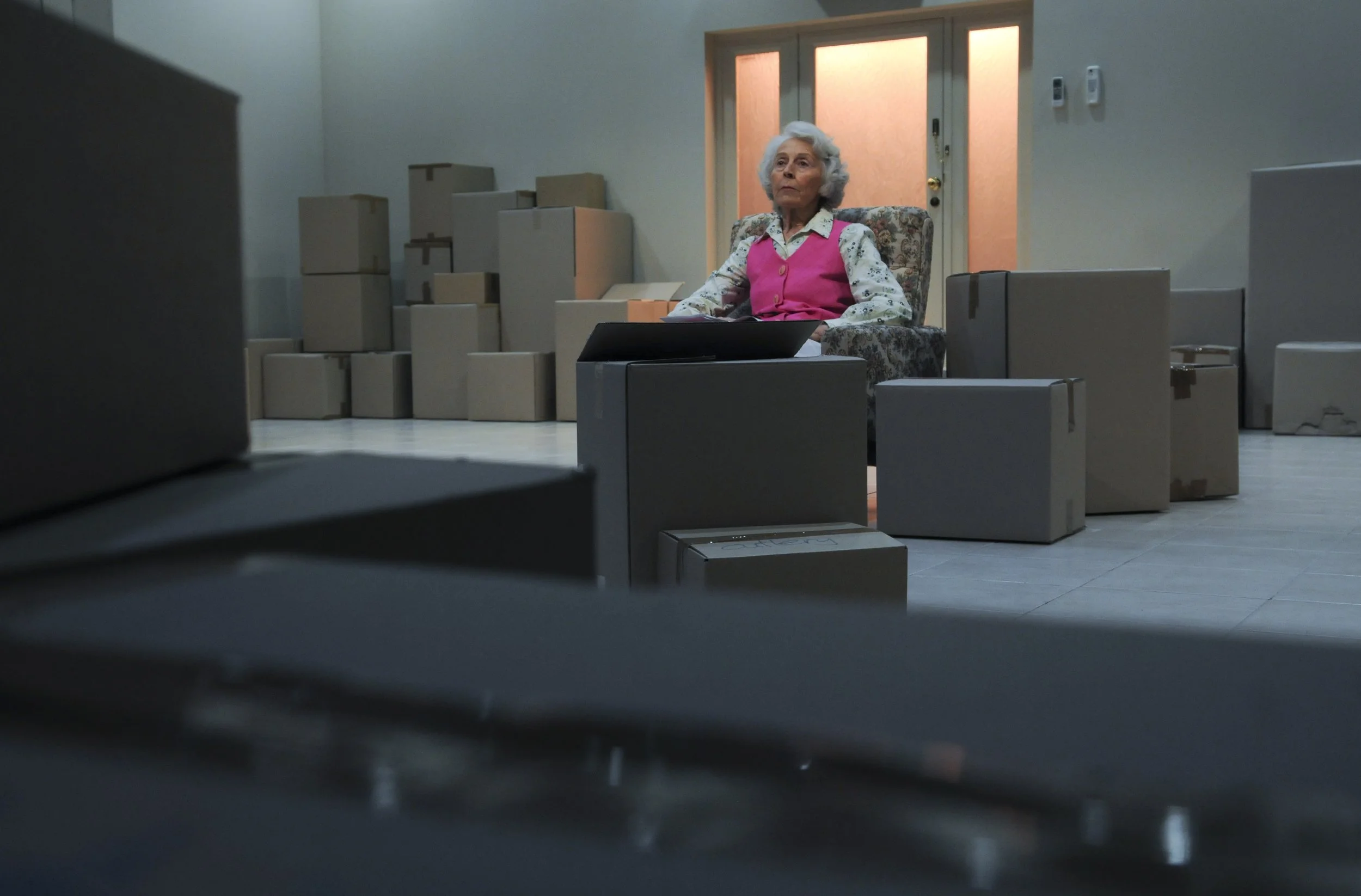 An elderly woman with white hair sitting in a floral armchair surrounded by numerous cardboard boxes in a room with a closed door and a light on the ceiling.