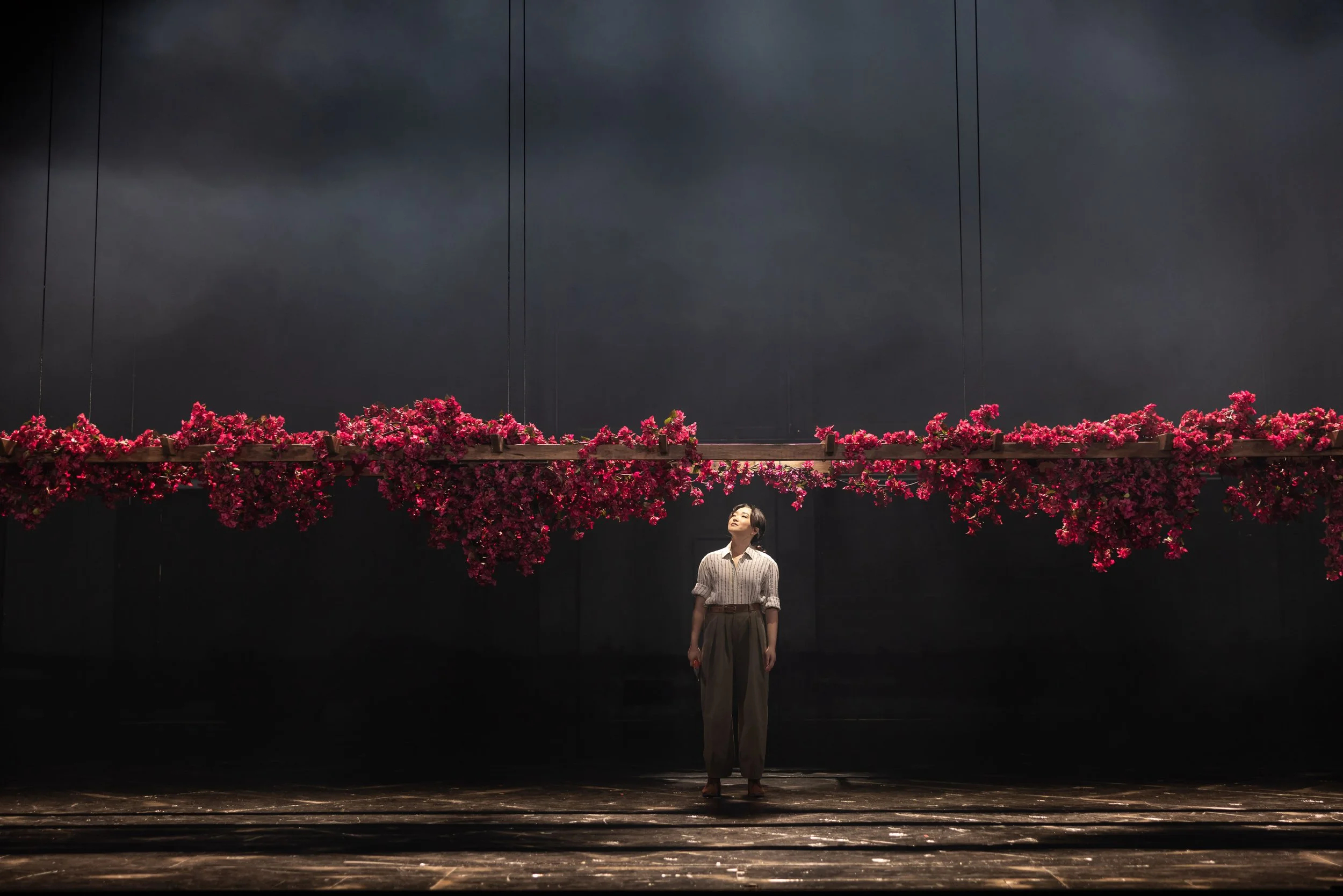 A woman standing under a large horizontal floral arrangement of pink flowers, in front of a dark background with stage lights.