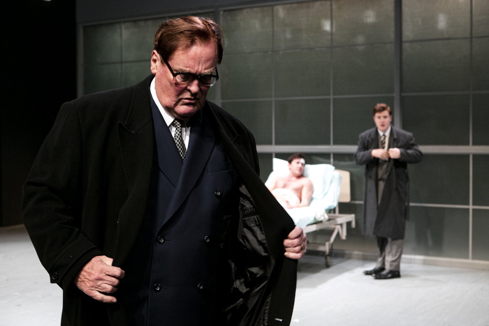An older man with glasses and brown hair looking down, in a dark coat, standing in front of a hospital bed with a woman lying in it, and a young man in a school uniform in the background