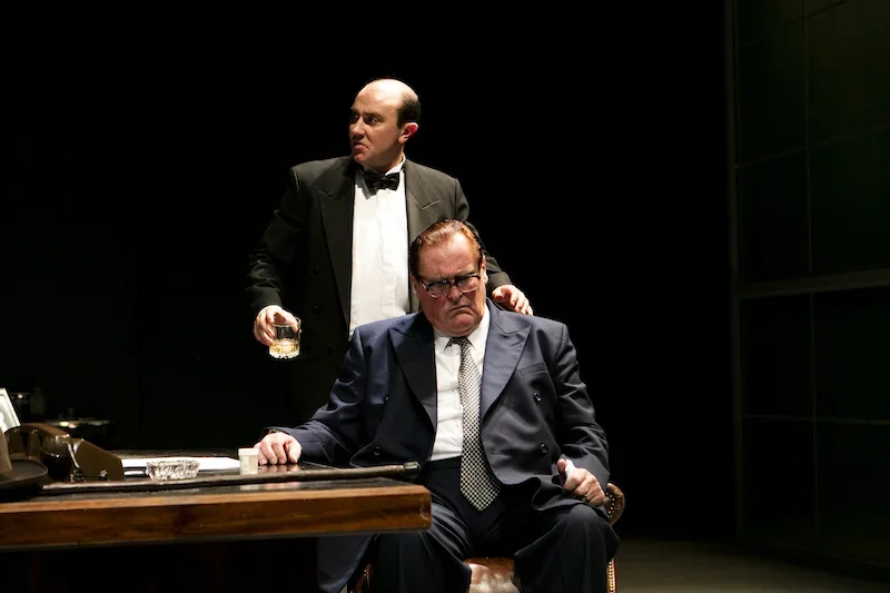 Two men in suits on stage, one sitting at a desk and the other standing behind him with a glass of whiskey, in a theatrical performance setting.