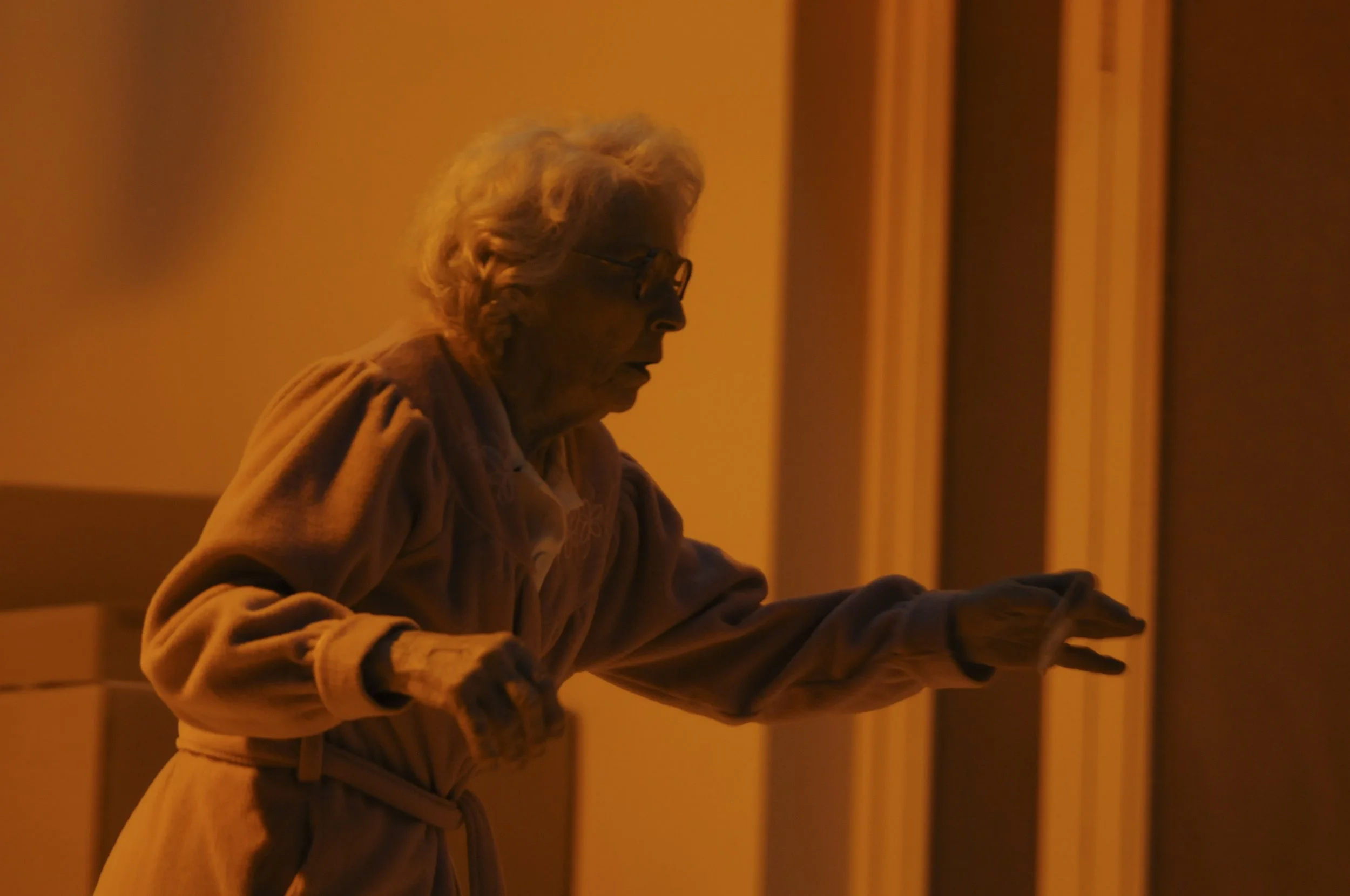 An elderly woman with glasses, wearing a jacket, sits at a table with her head slightly bowed, in a dimly lit room with an orange tint.