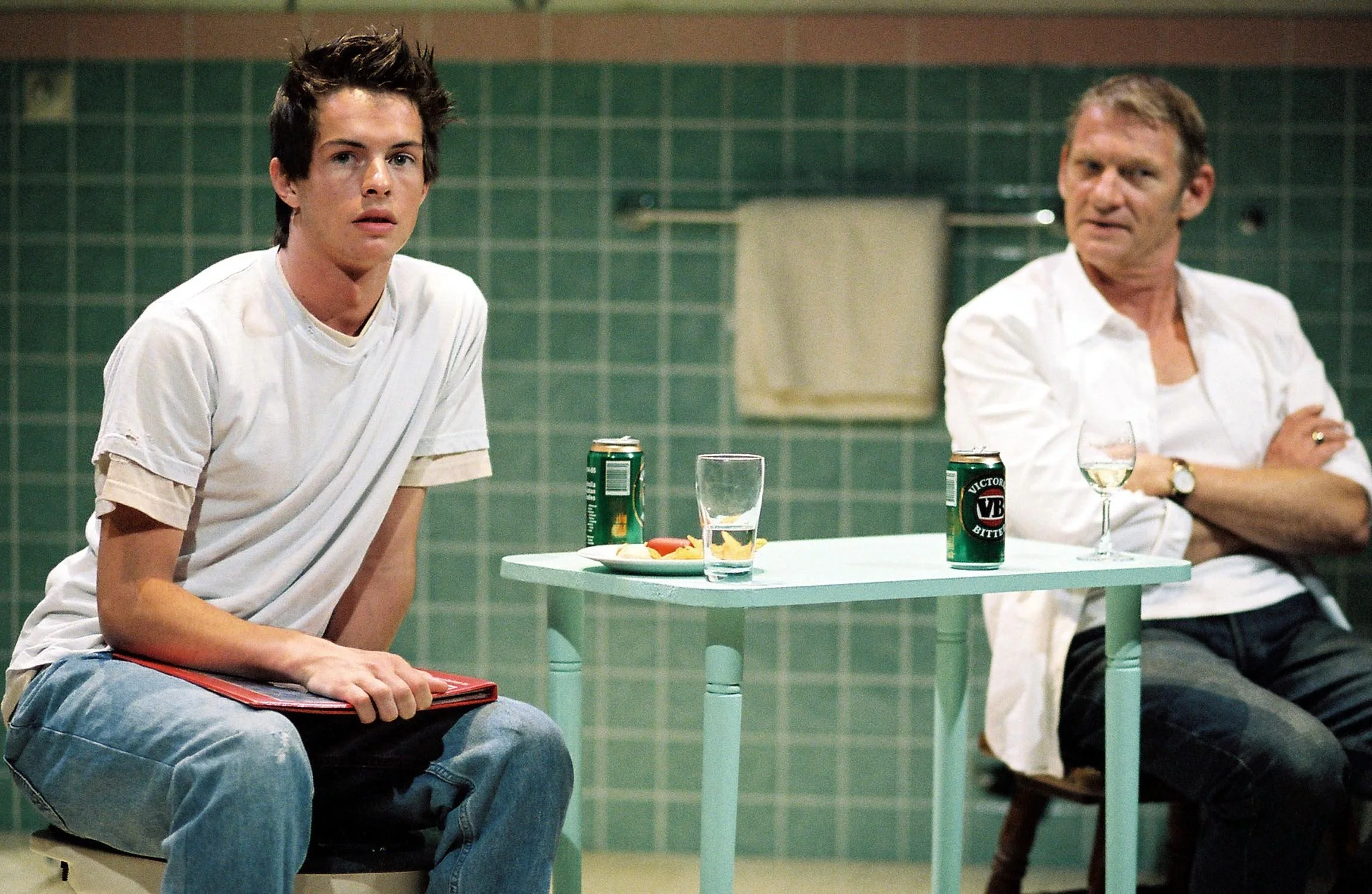 A young man with dark, spiked hair, wearing a white t-shirt and ripped jeans, seated at a table in a room with green tiled walls. He has a serious expression. On the table, there is a plate of food, a can of VB beer, a glass of water, and a napkin. A