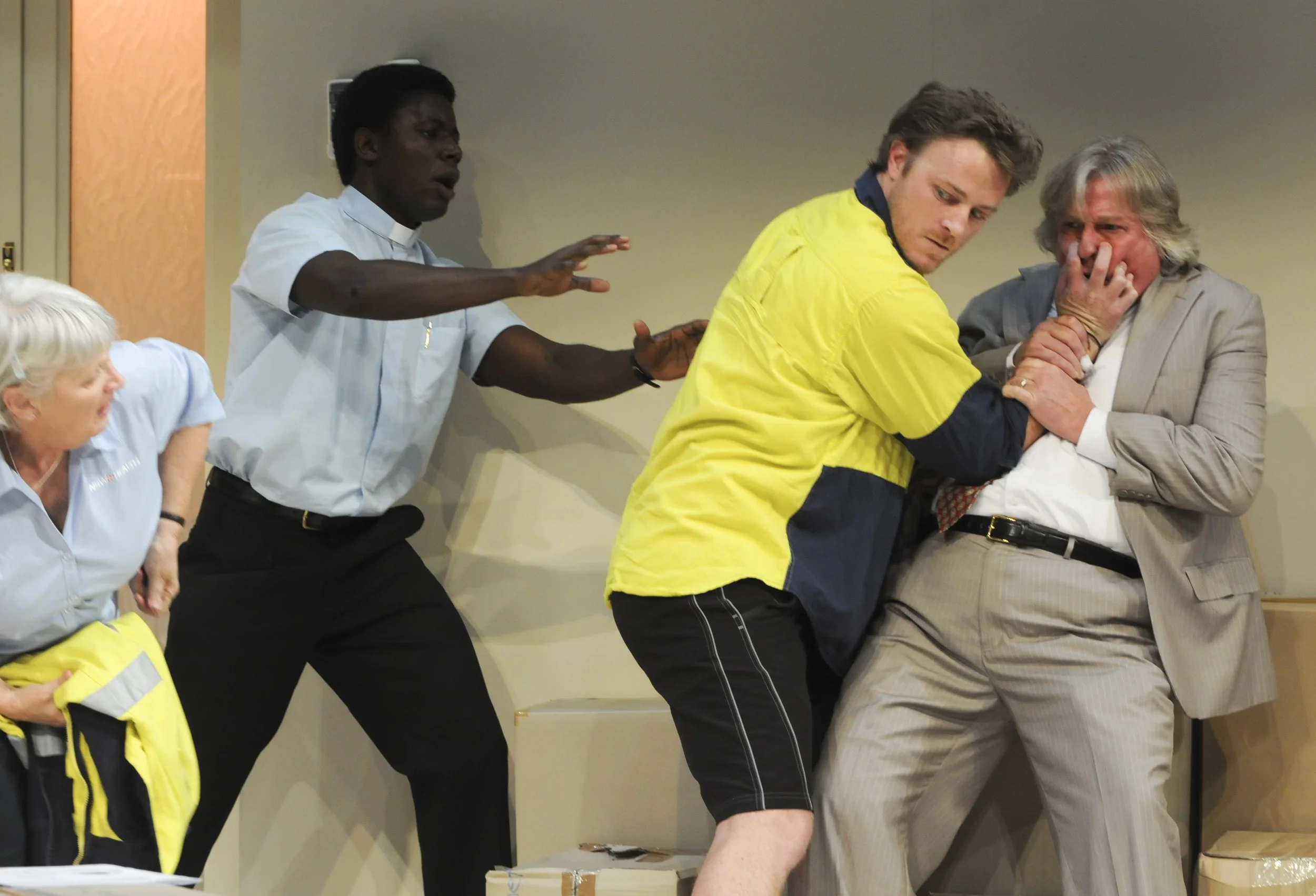 A group of people participating in a staged mock fight, with a man in a yellow shirt holding another man in a gray suit in a headlock, while others watch and react with alarm or concern.