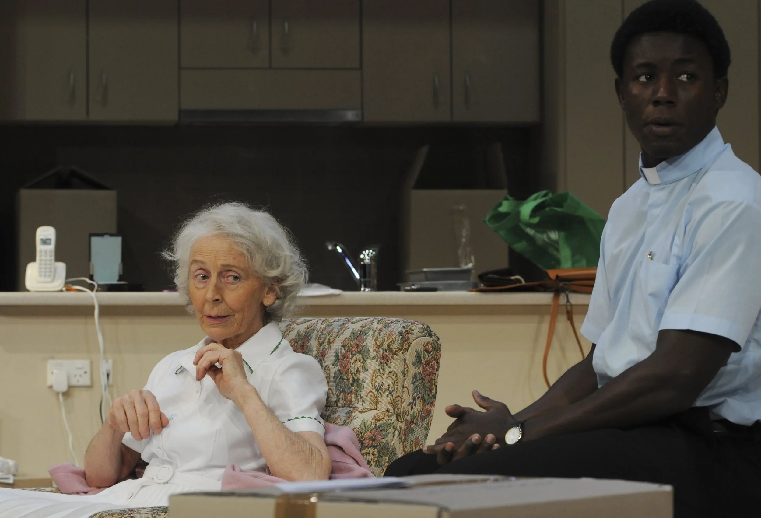 An elderly woman with white hair sitting in a hospital bed and a young man in a nurse's uniform standing beside her in a hospital room.
