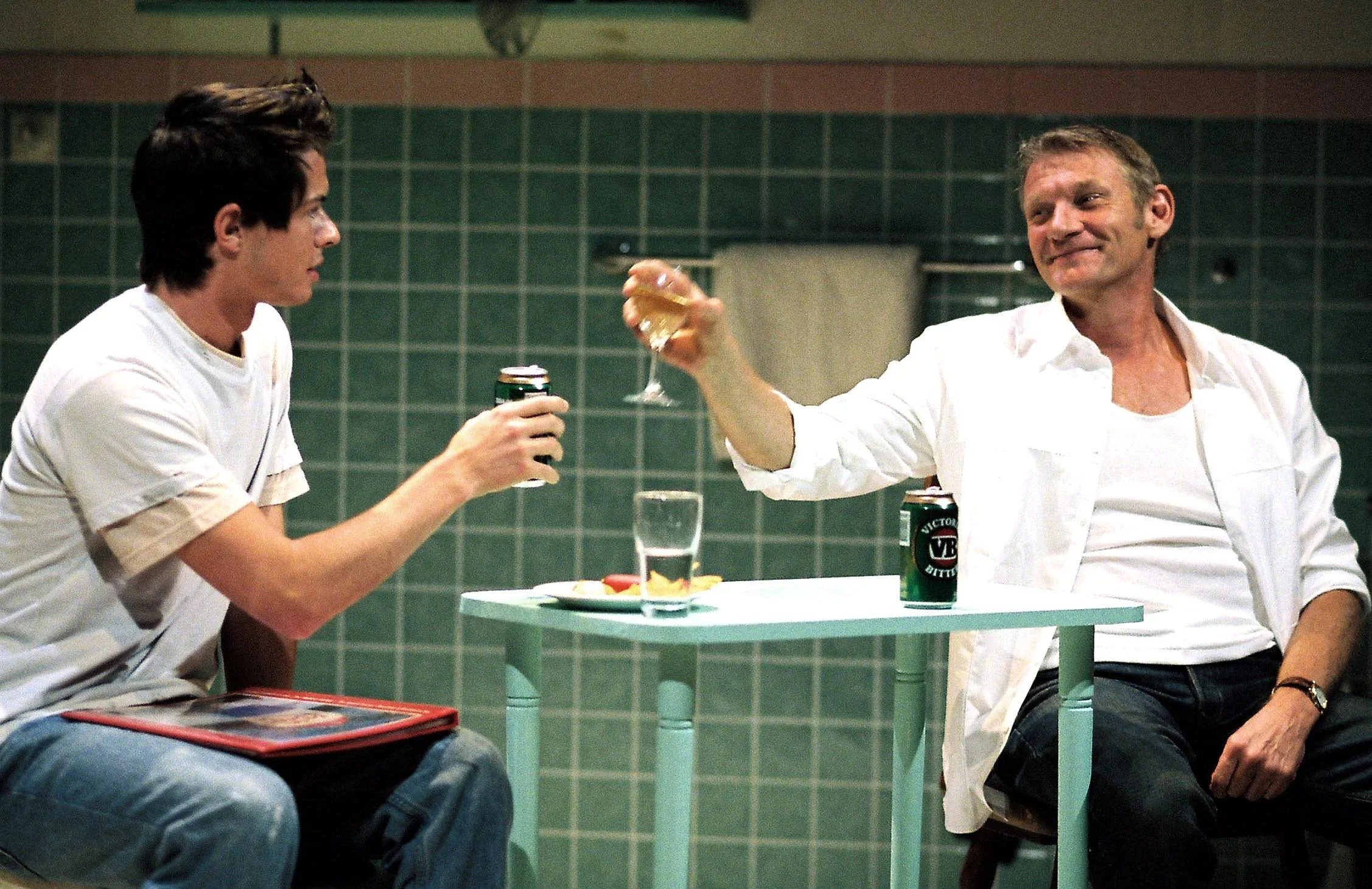 Two men sitting at a small round table with drinks. One man is holding a can and the other is holding a glass, clinking glasses in a toast, with a green tiled wall in the background.