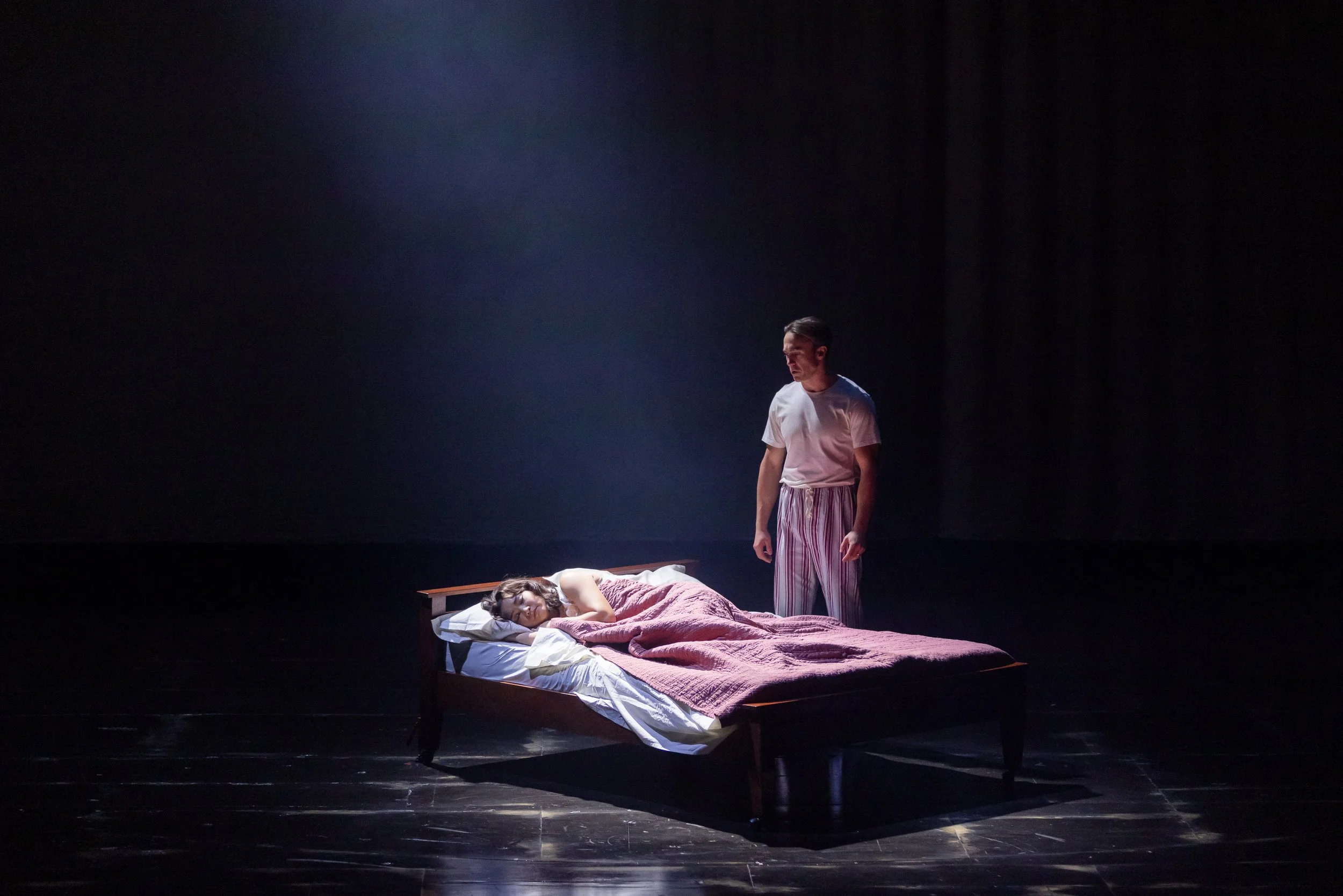 A stage scene with a woman lying on a bed covered with a pink blanket, and a man standing beside her on a dark stage with minimal lighting.