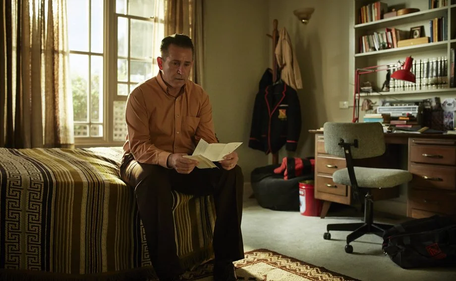 A man in a tan shirt sitting on a bed, reading a book, in a bedroom with sunlight coming through the window, a desk with a red lamp, a gray chair, a bookshelf, and a coat hanging on the wall.