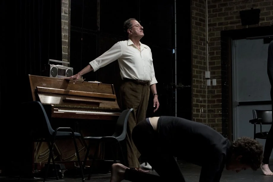 A person with glasses and gray hair, wearing a white shirt and brown pants, standing on a stage beside a piano, with a black brick wall in the background. Another person is kneeling and bending forward in the foreground.