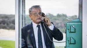 Man in suit and glasses talking on a landline phone, seen through a glass window, with a teal briefcase nearby.