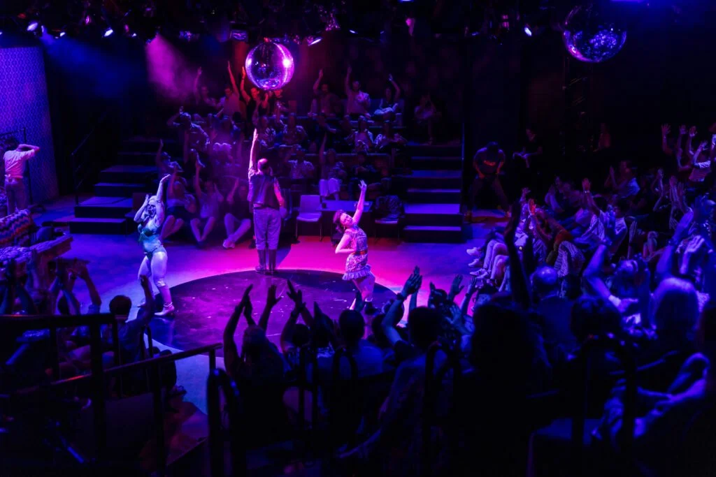 A circus or dance performance with performers on a stage surrounded by an audience, with purple lighting and disco balls overhead.