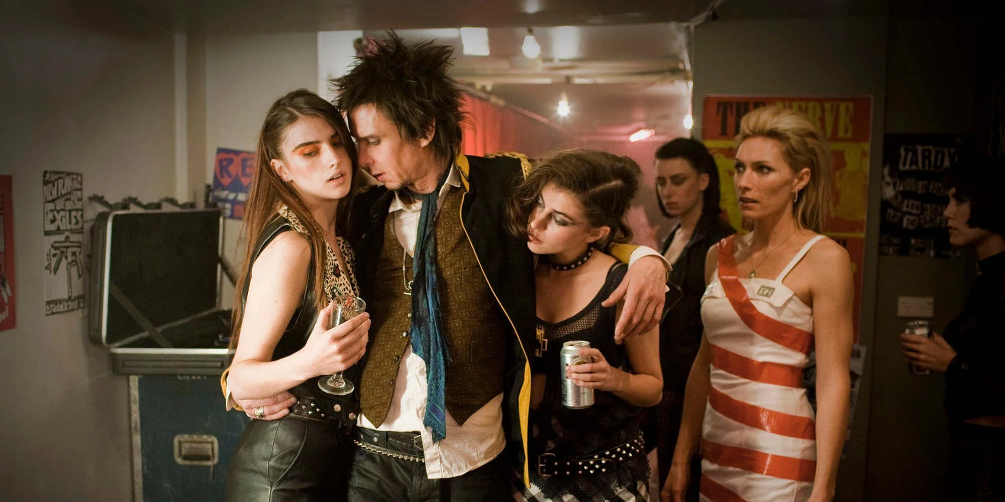 Group of five young adults at a party, some holding drinks, with graffiti and posters in the background.