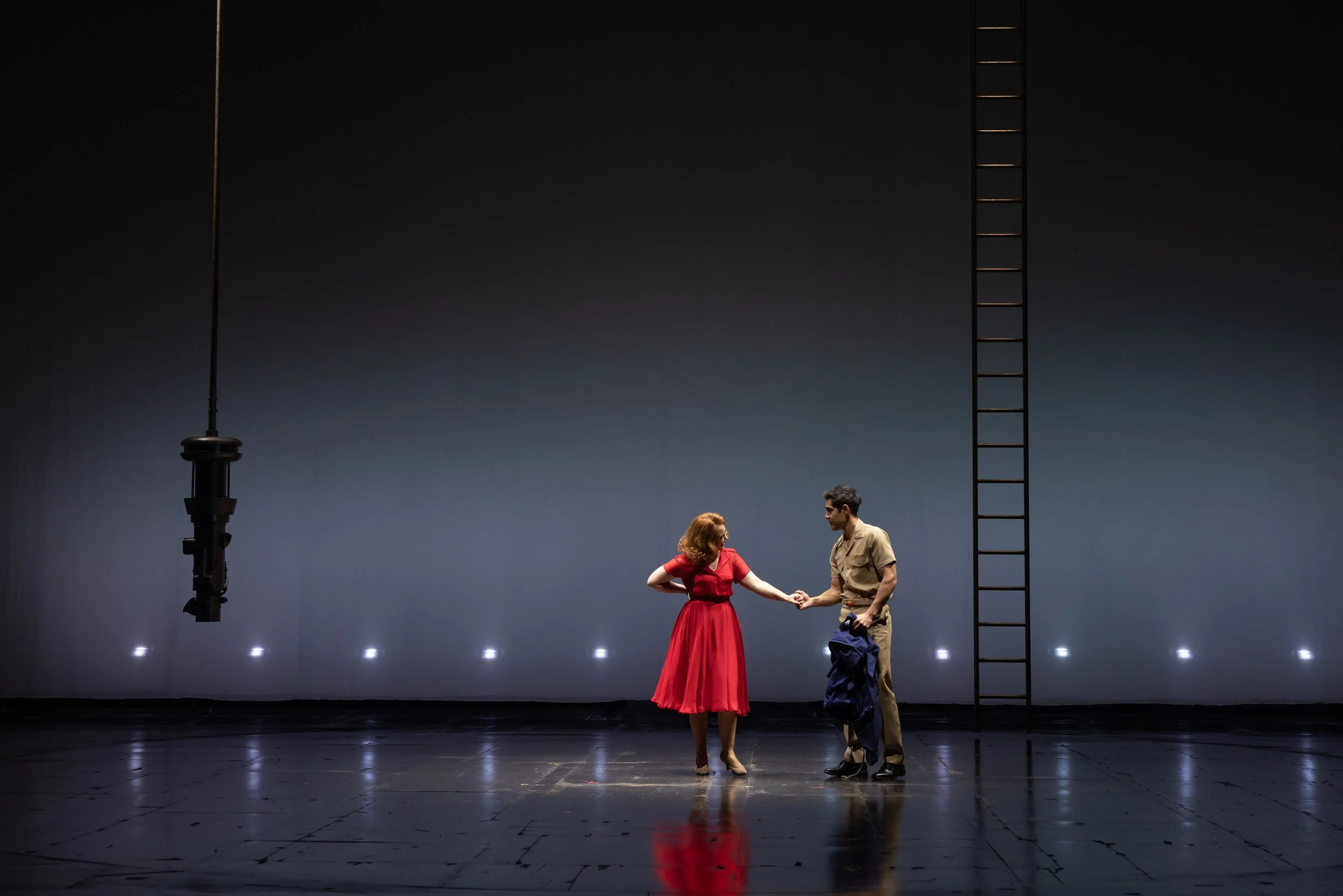 A woman in a red dress and a man in a beige outfit hold hands on a stage, with a large ladder and stage lights in the background.