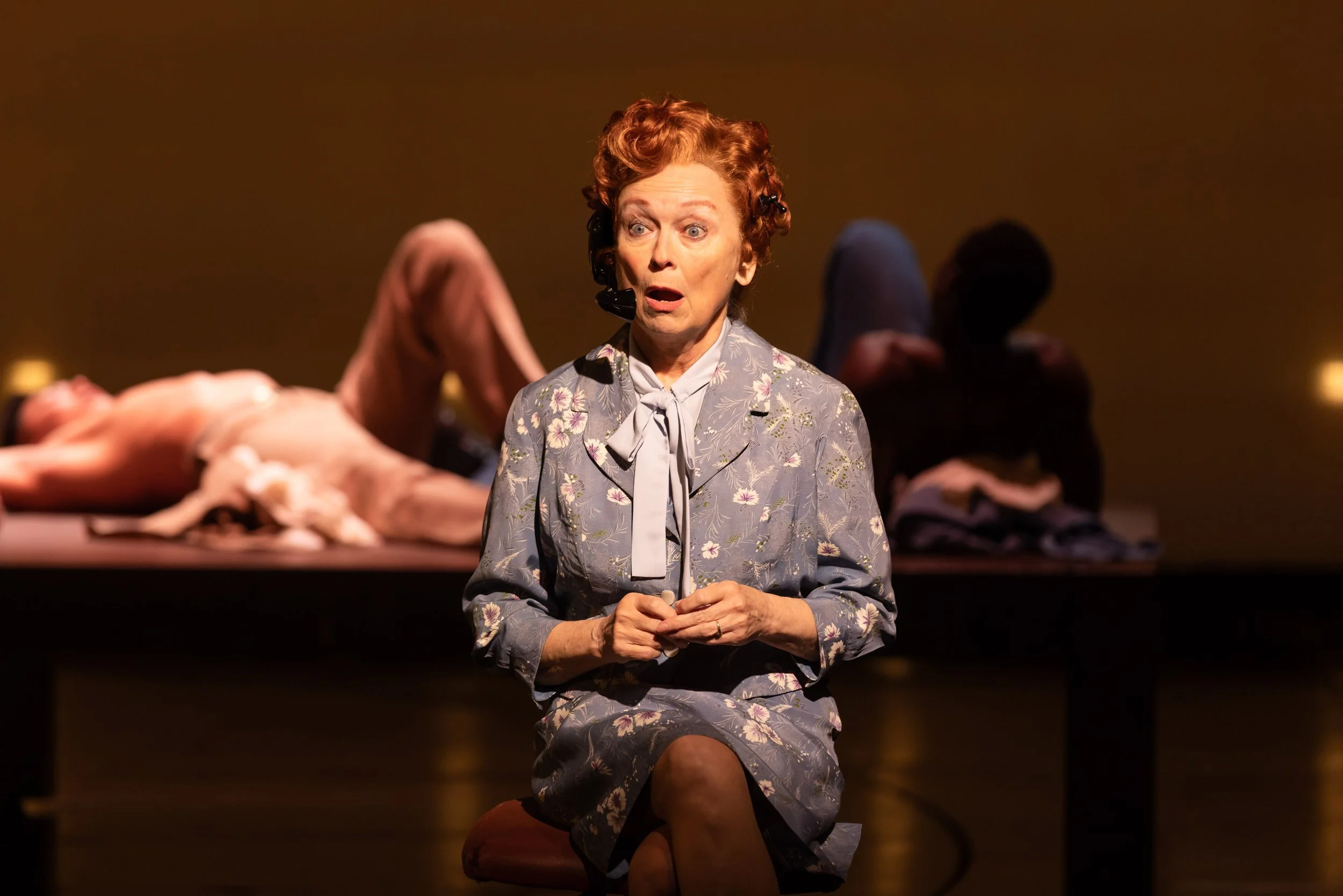 An elderly woman with red curly hair, wearing a floral gray blouse with a neck bow, sits with her hands clasped, looking surprised or shocked. Behind her are several mannequins on display, dressed in vintage clothing, in a dimly lit setting.