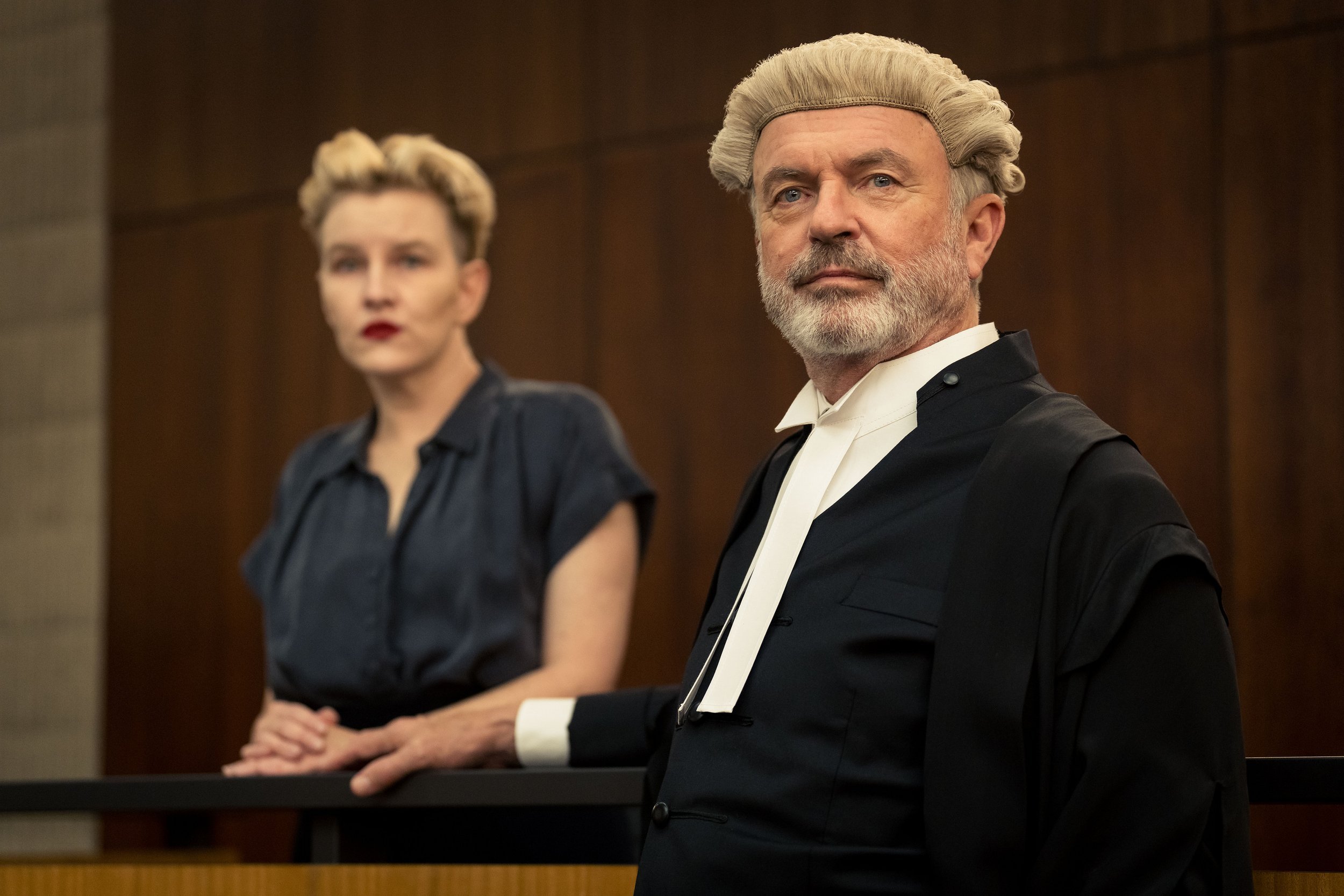 A judge with gray hair, dressed in a black robe with a white collar, stands with a serious expression in a courtroom. A woman with blonde hair and a black top is blurred in the background, also standing in the courtroom.