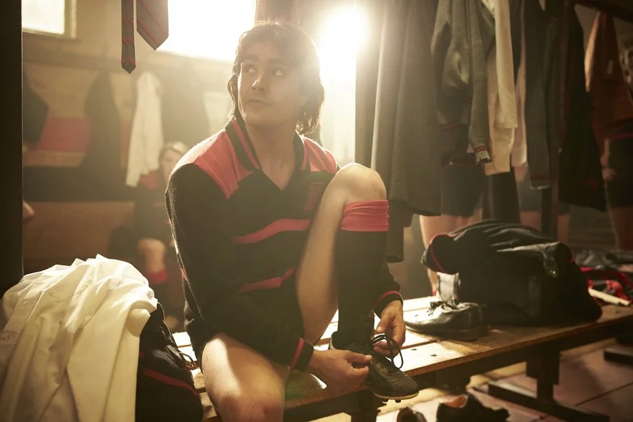A person sitting on a bench in a locker room, putting on a shoe, with sports clothing and bags around, and sunlight coming through the window.