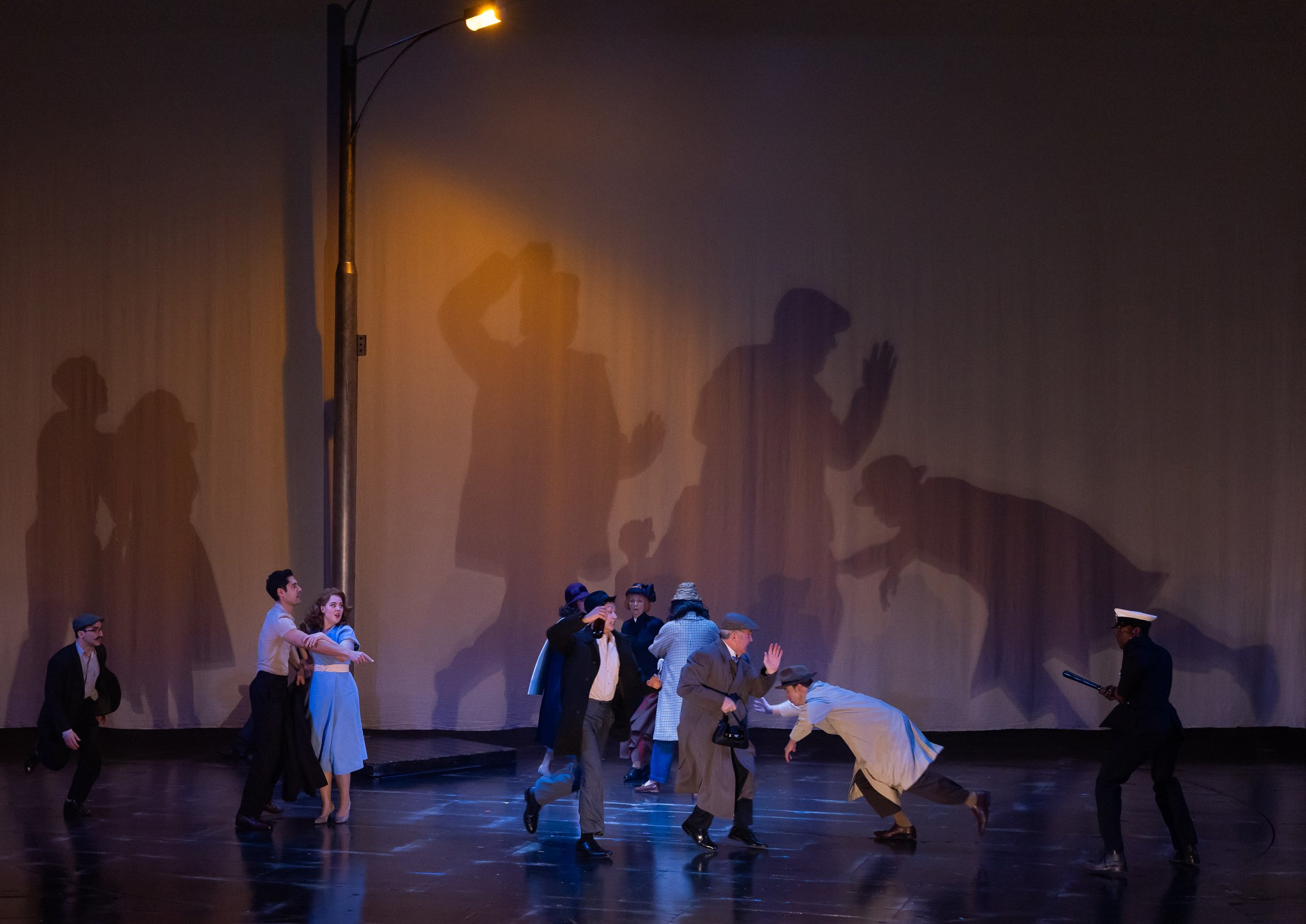The image shows a theatrical stage performance with actors in period costumes. Some actors are standing, while others are crouched or bending in a choreographed scene. The backdrop is a plain wall with dramatic shadows of the actors projected onto it