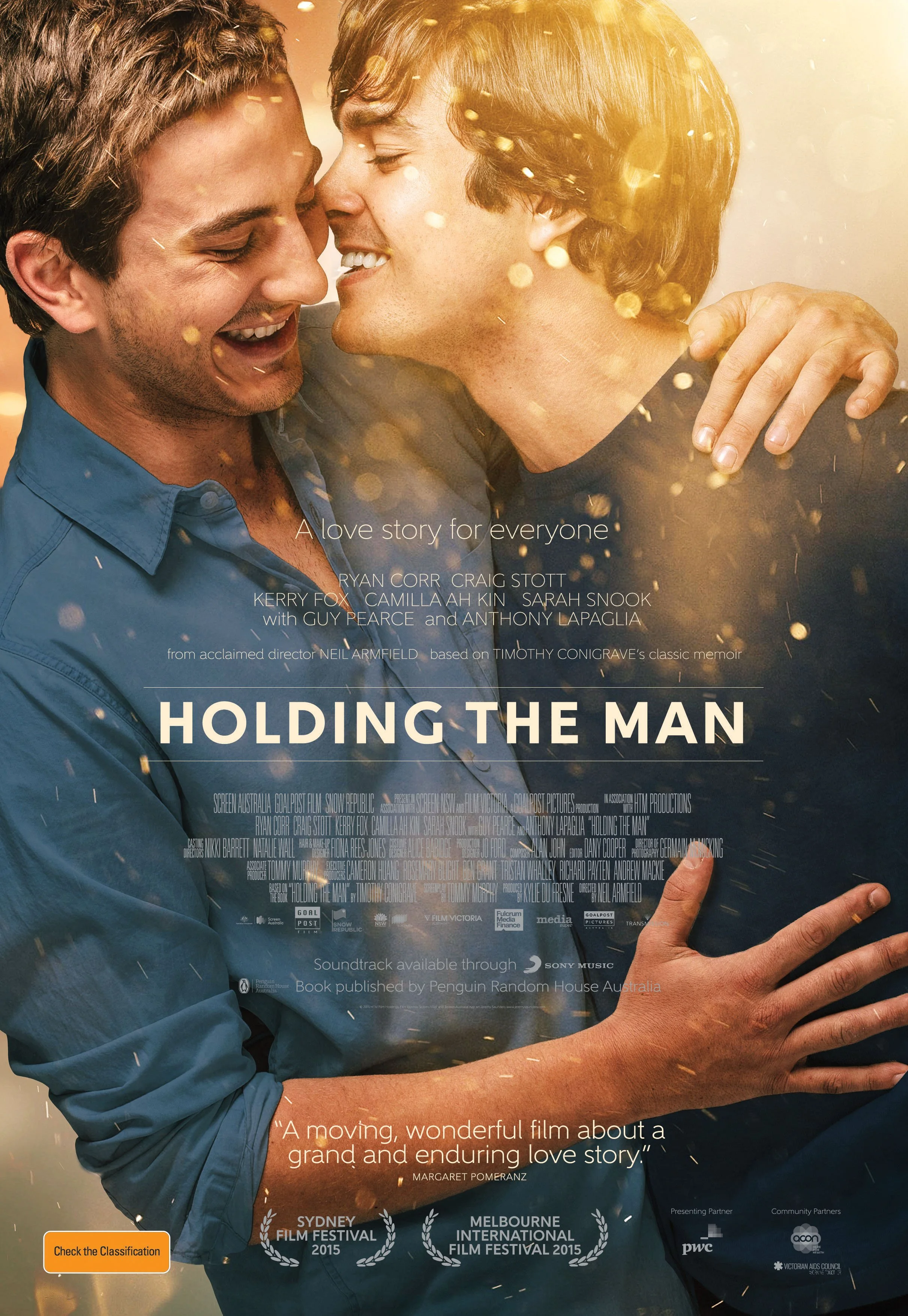Close-up of two men, one with darker hair and the other with longer lighter hair, smiling and embracing each other. The man with lighter hair has his hand on the other's chest, and they appear to be about to kiss. Golden light and bokeh effects creat