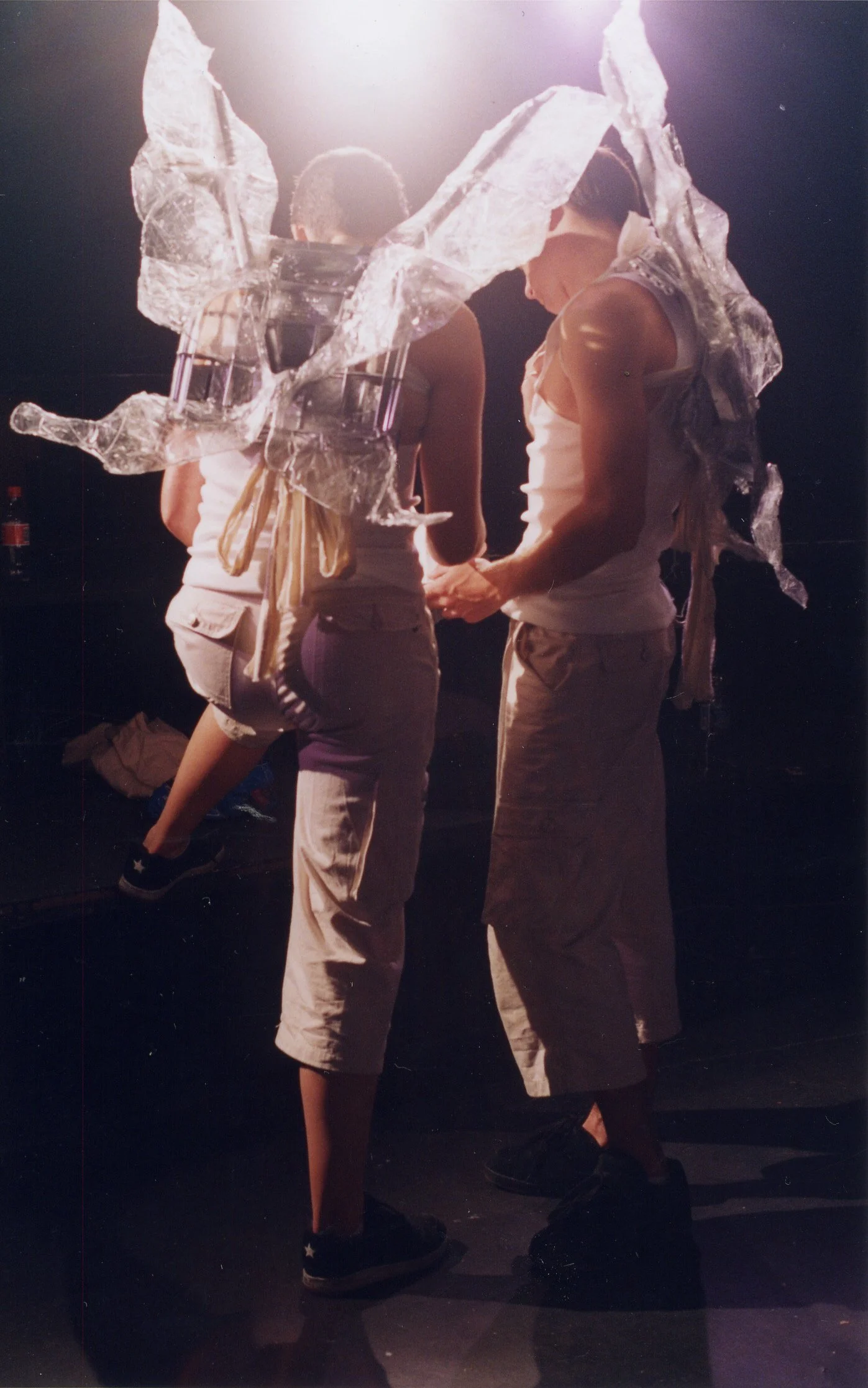 Two people with shaved hair wearing white tank tops and beige pants, standing close together in a dark room illuminated by bright light overhead. They are facing away or sideward, and one person appears to be holding a mobile phone. A large, abstract sculptural piece made of translucent material is held above them.