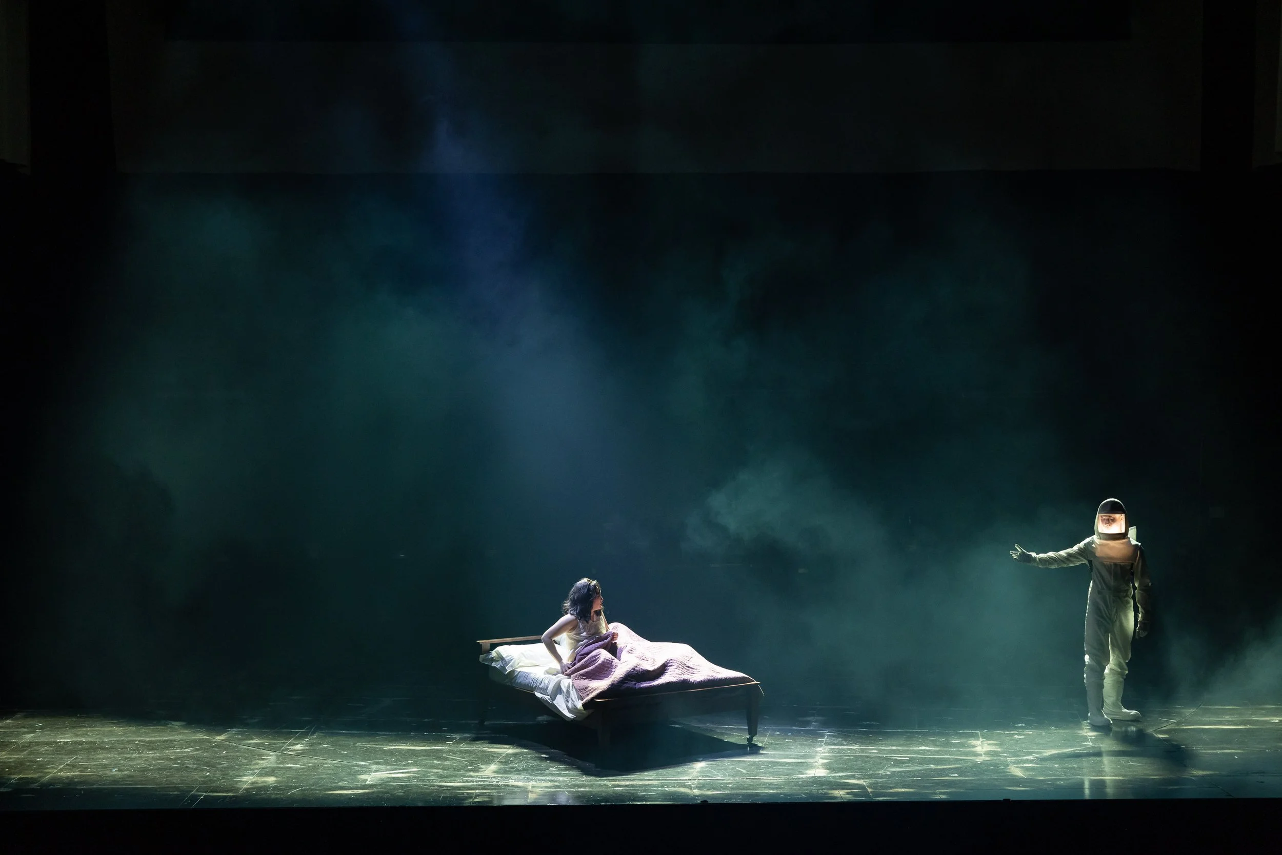 A theatrical scene with a woman lying on a bed and a person in a hazmat suit standing nearby, set against a dark background with stage lighting.