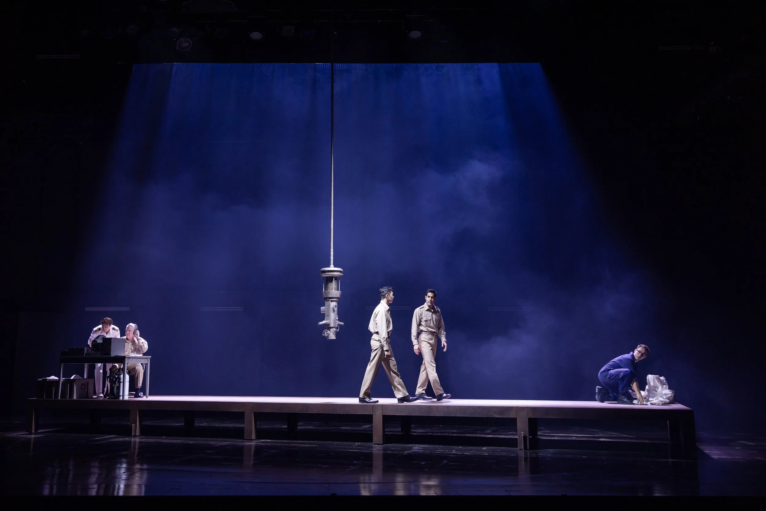 A stage scene with six people dressed in vintage military or scientific uniforms, some sitting at a table working on a computer, others standing or bending down, with a large tall antenna-like structure in the center, and a blue curtain with textured