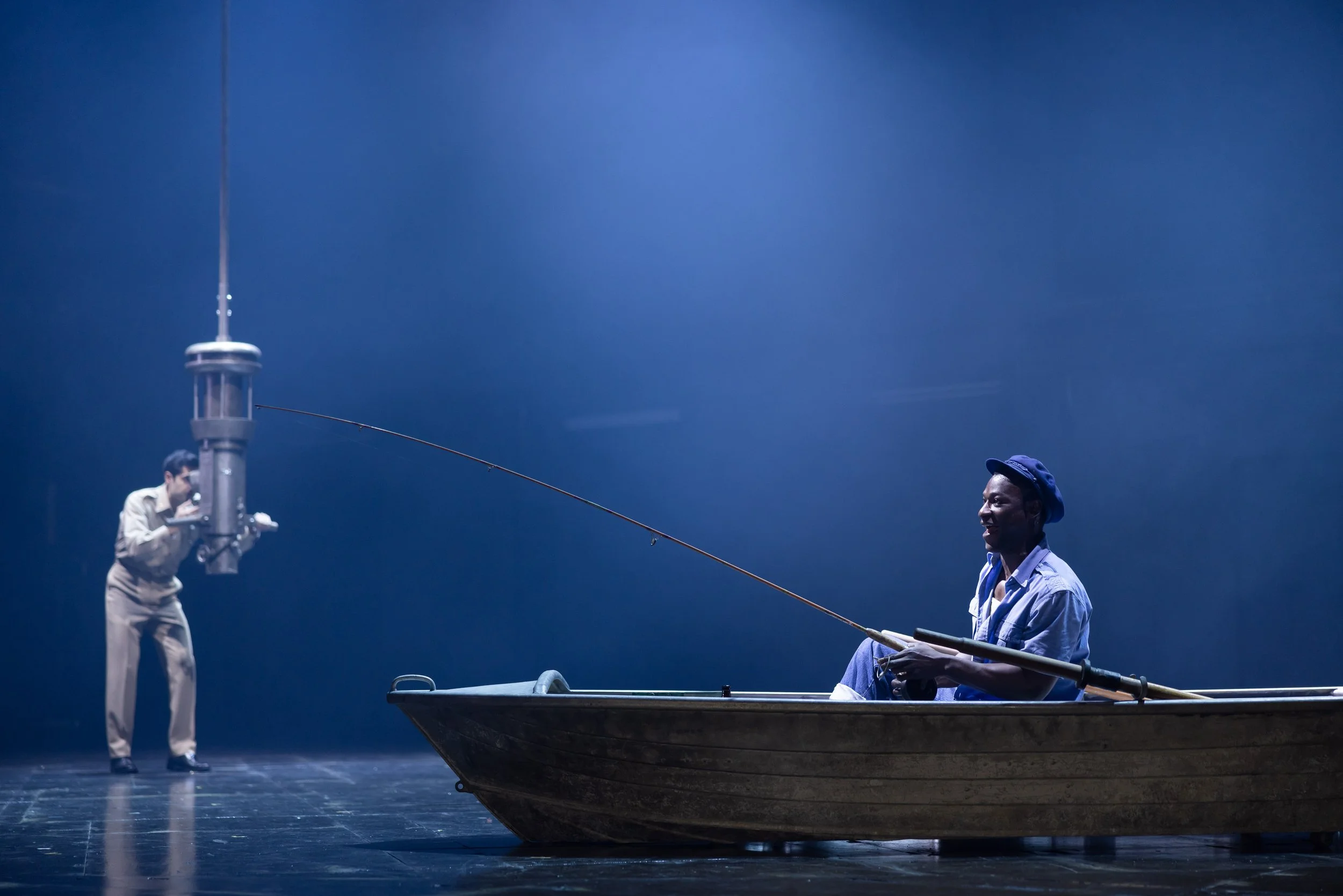 Two men on a stage with a small boat, one man is fishing with a pole and the other is dressed casually, standing nearby and smiling, with a large blue backdrop.