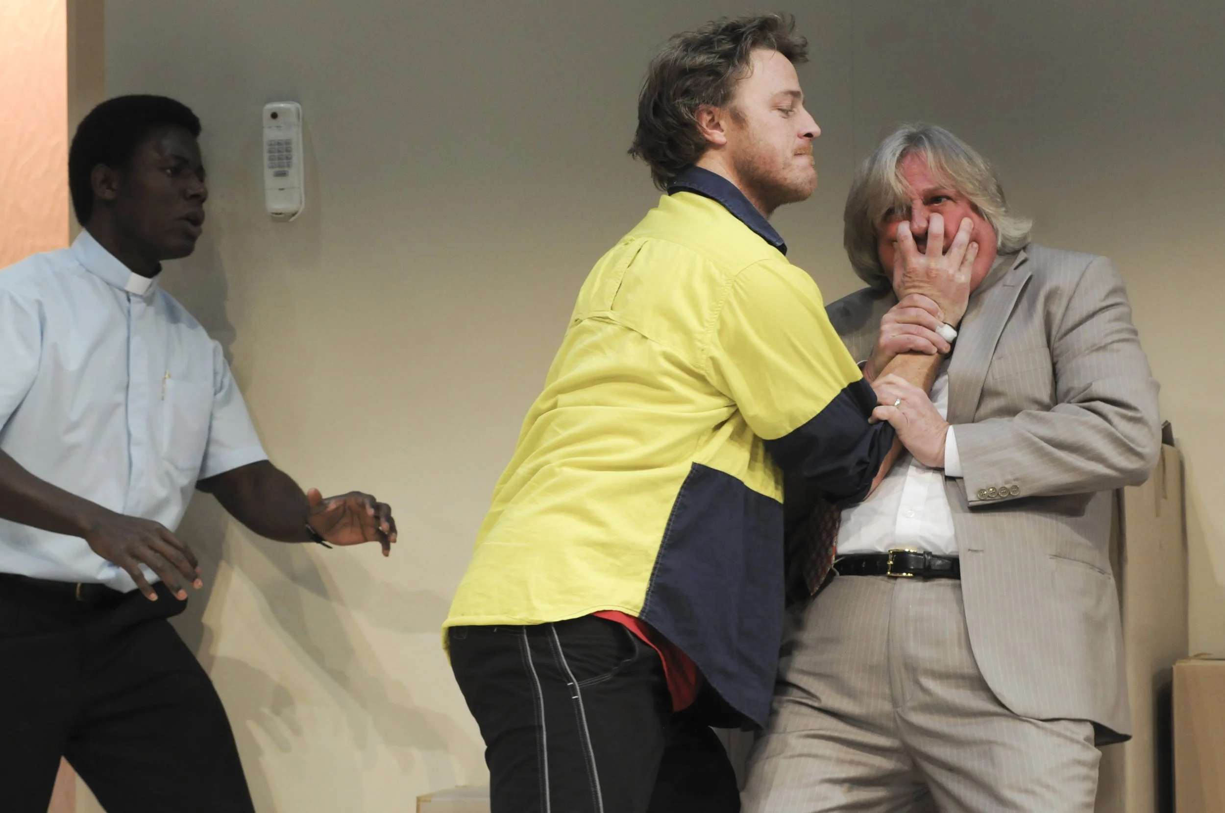 A scene inside a courtroom or meeting room featuring three individuals. One appears to be a priest or religious figure on the left, dressed in a white clerical shirt. The middle individual, a young man with light brown hair, is wearing a yellow jacke