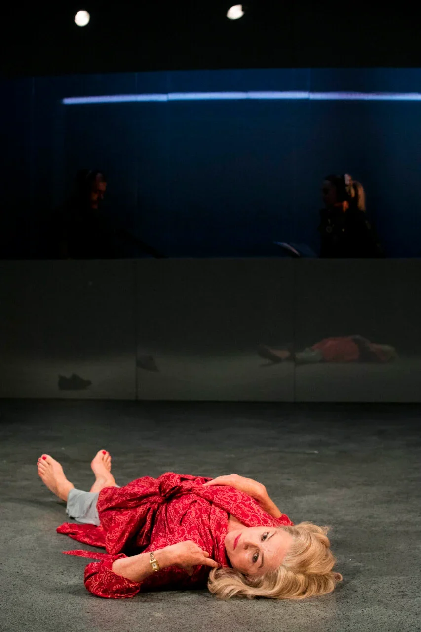 A woman with blonde hair lying on the floor on her side, wearing a red patterned dress, with a neutral expression, in a dark room with a reflective black surface behind her and two people in the background taking photos.