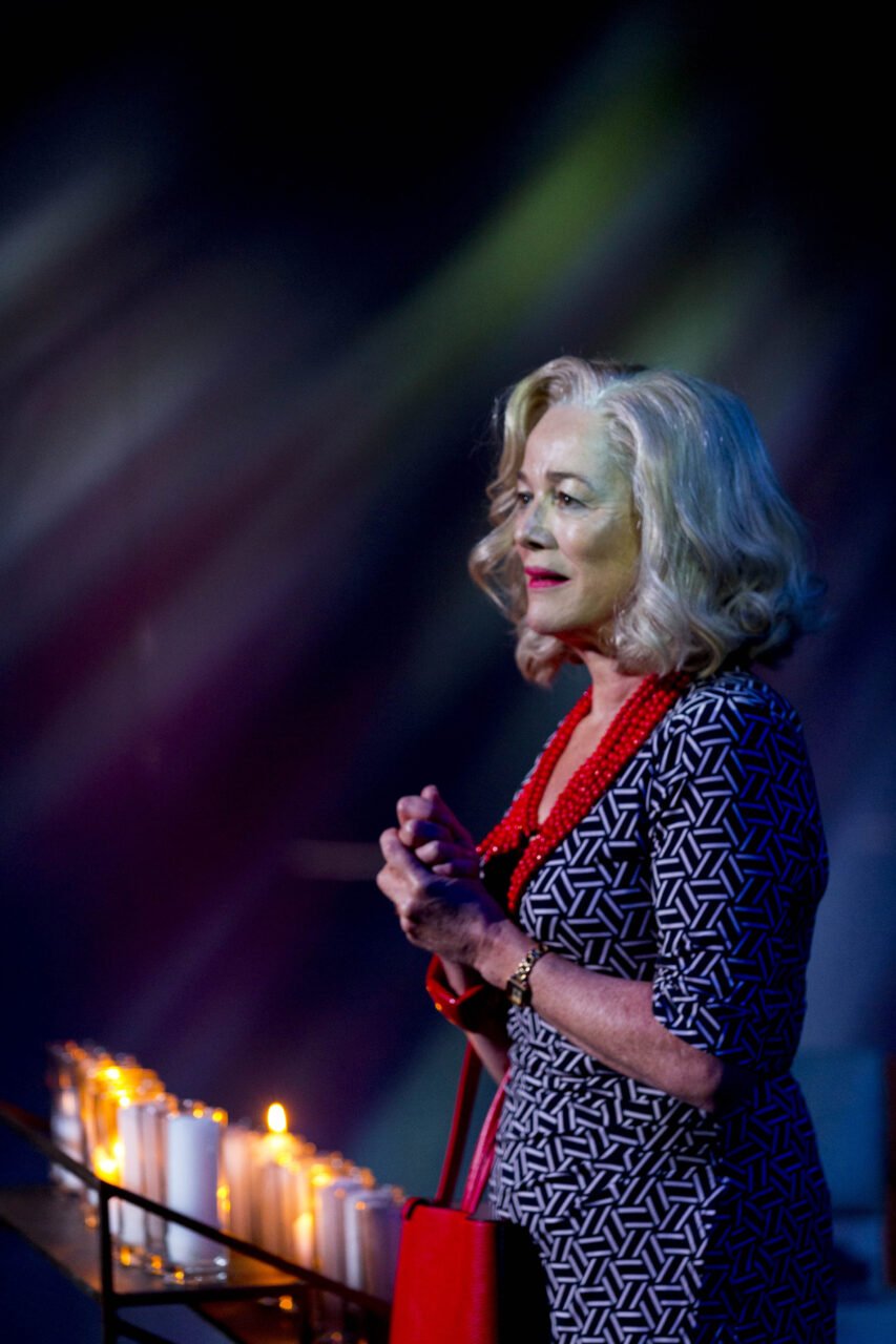 A woman with grey wavy hair and light skin stands with her hands clasped, illuminated by candles, wearing a patterned dress, red necklace, and a red purse, in a dimly lit setting.