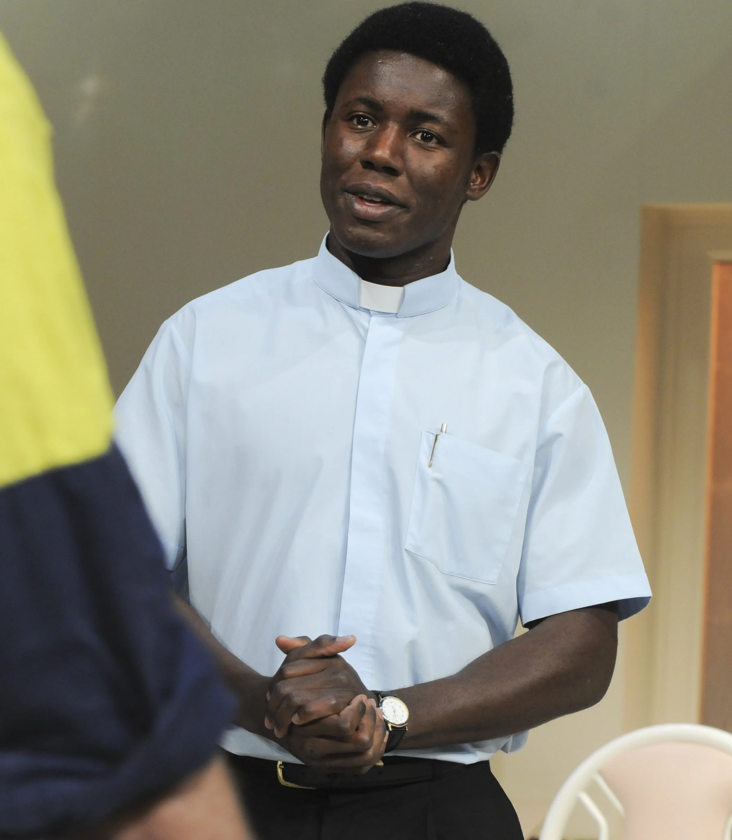 A man with dark skin, wearing a light blue clerical shirt, standing indoors, and clasping his hands together in front of him. He is wearing a watch on his left wrist.