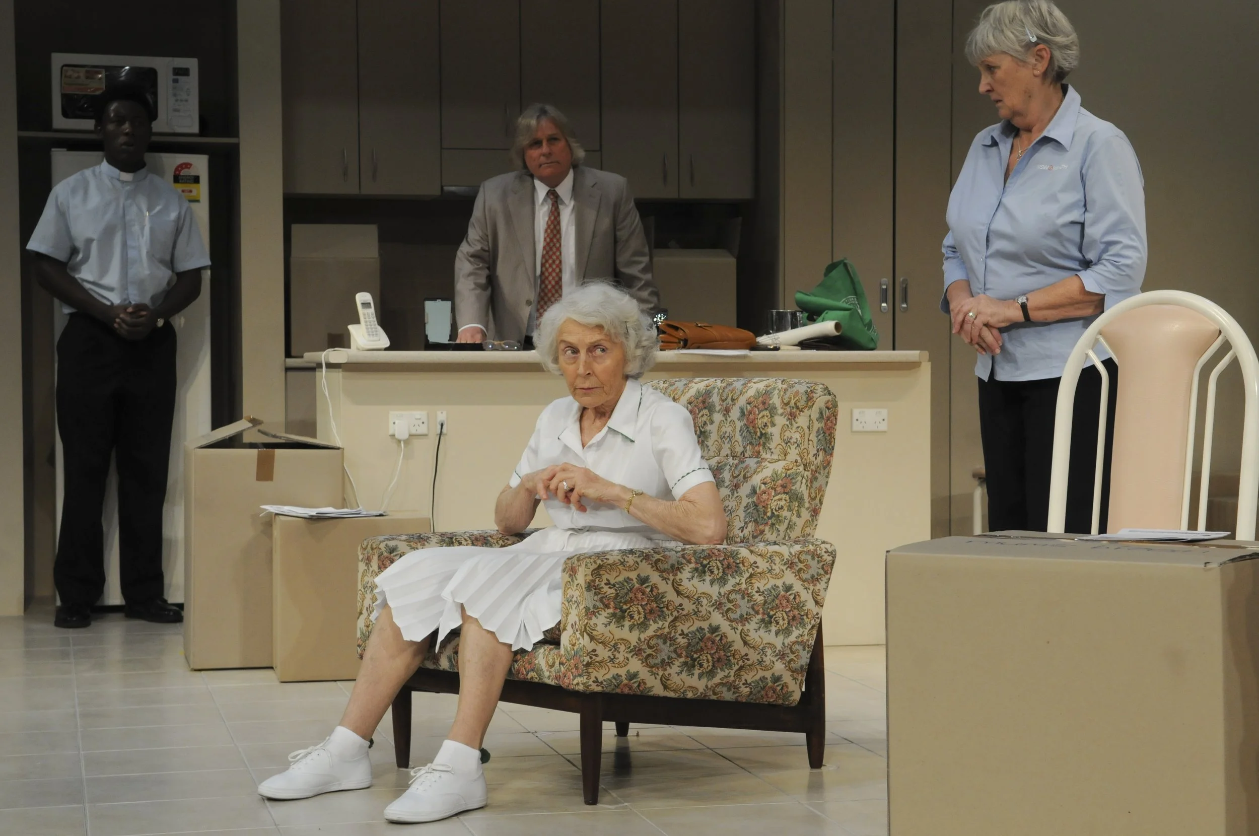 An elderly woman with white hair sitting on a floral patterned chair with her legs crossed, wearing a white dress and white sneakers, appears to be in a nursing home or hospital lounge. Behind her, four adults stand: a young woman in a blue shirt and