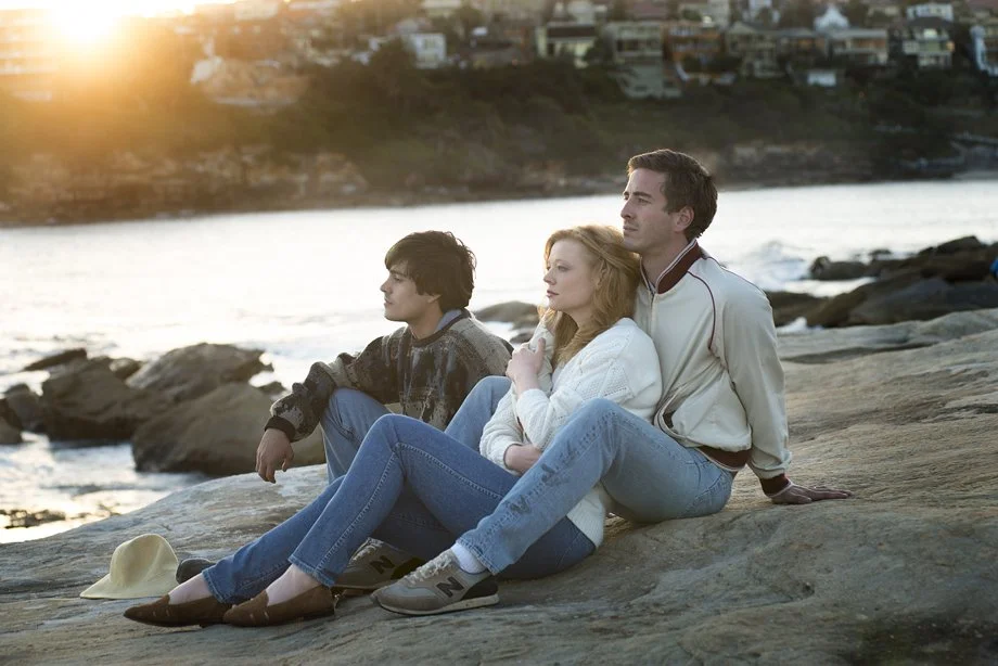 Three people sitting on rocks by the water at sunset, with a cityscape in the background.