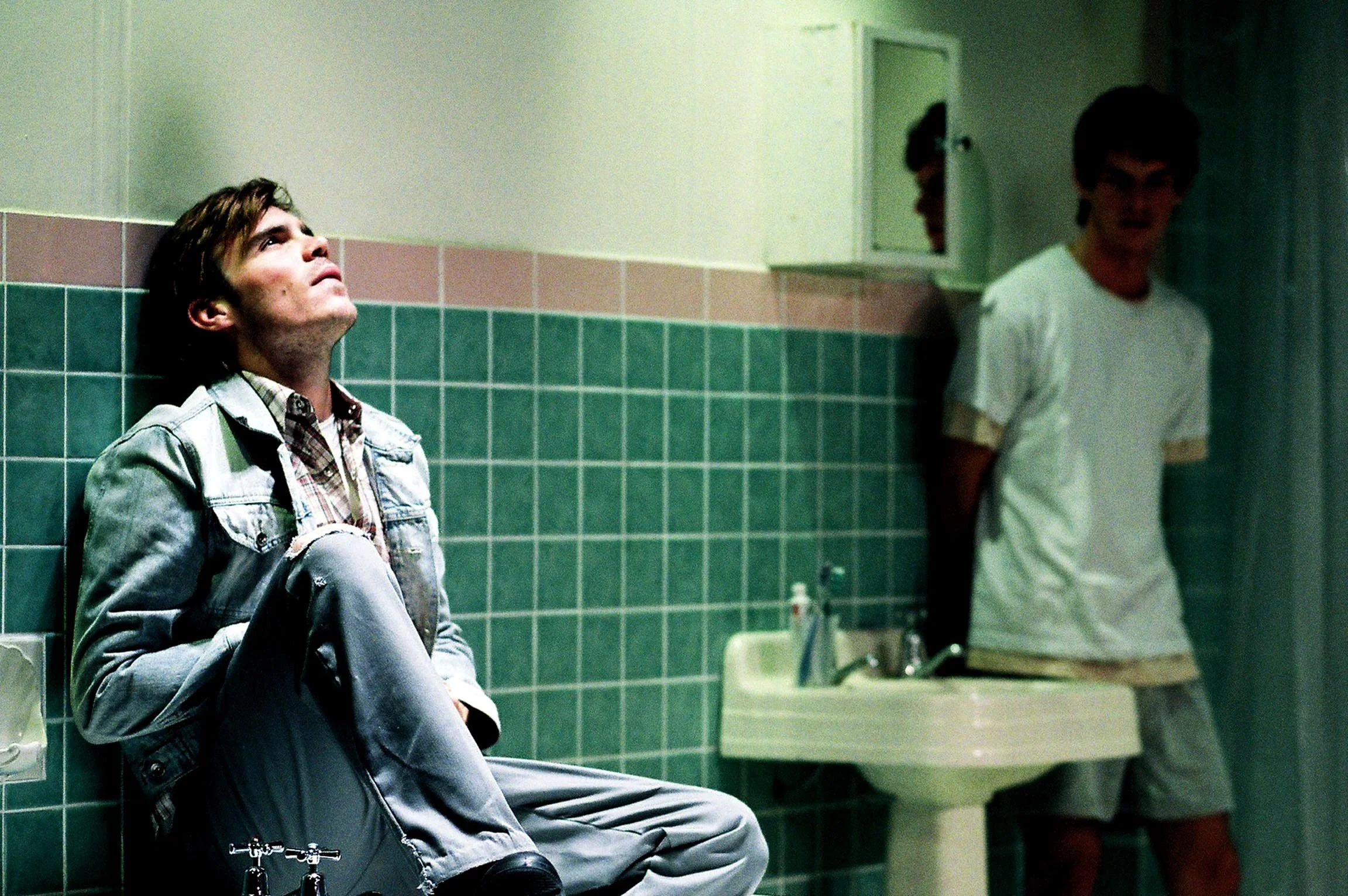 Young man sitting on tiled bathroom wall with head tilted back, looking up, in a restroom with mirror and sink, and another young man in the background.