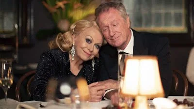 An elderly couple sitting at a dinner table, smiling and cuddling in a warmly lit restaurant.