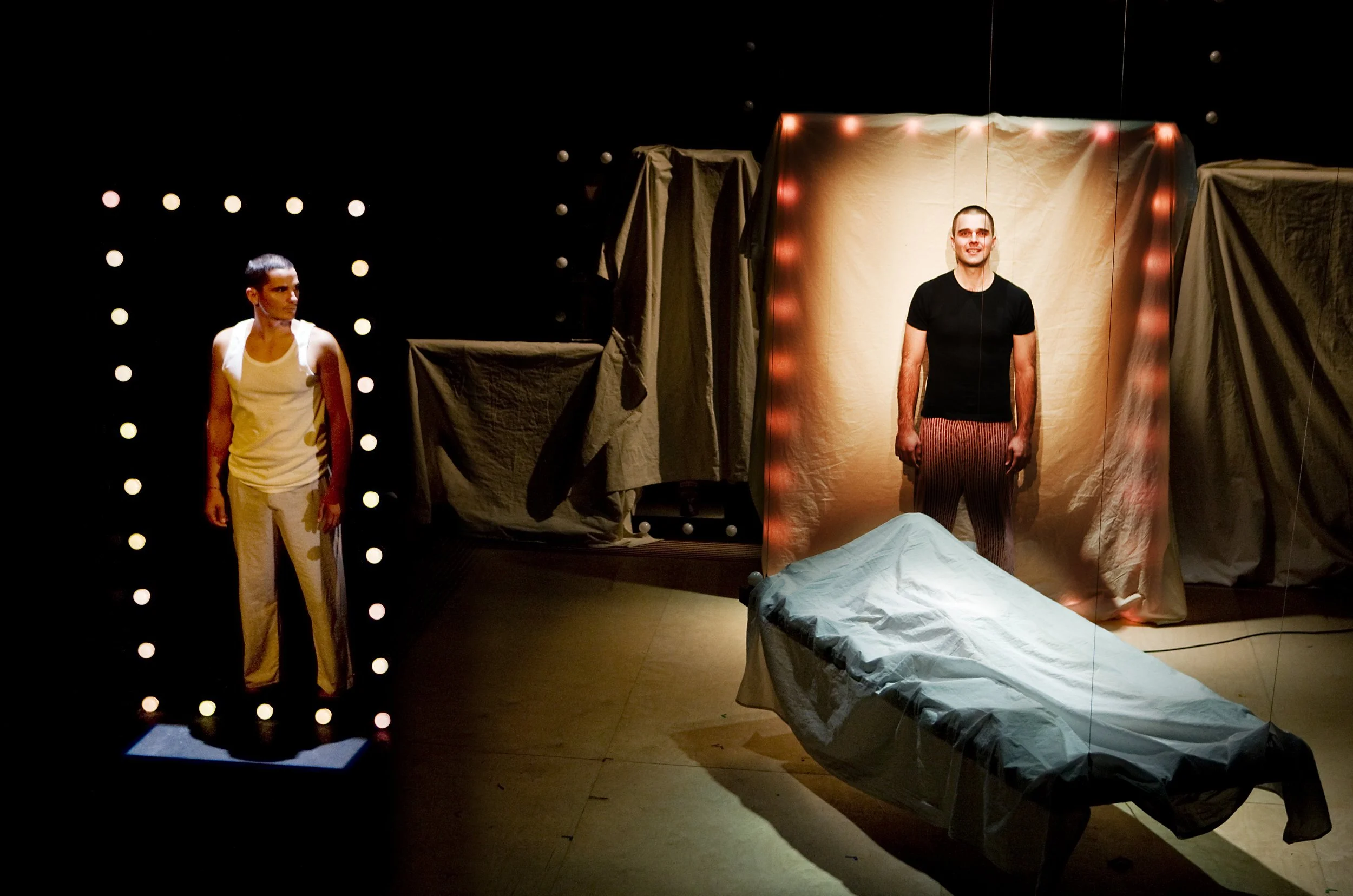 Two performers on stage with a backdrop of fabric and string lights. One person is on the left, wearing a white tank top and light-colored pants, and the other on the right, wearing a black shirt and striped pants. The stage is dimly lit with spotlig