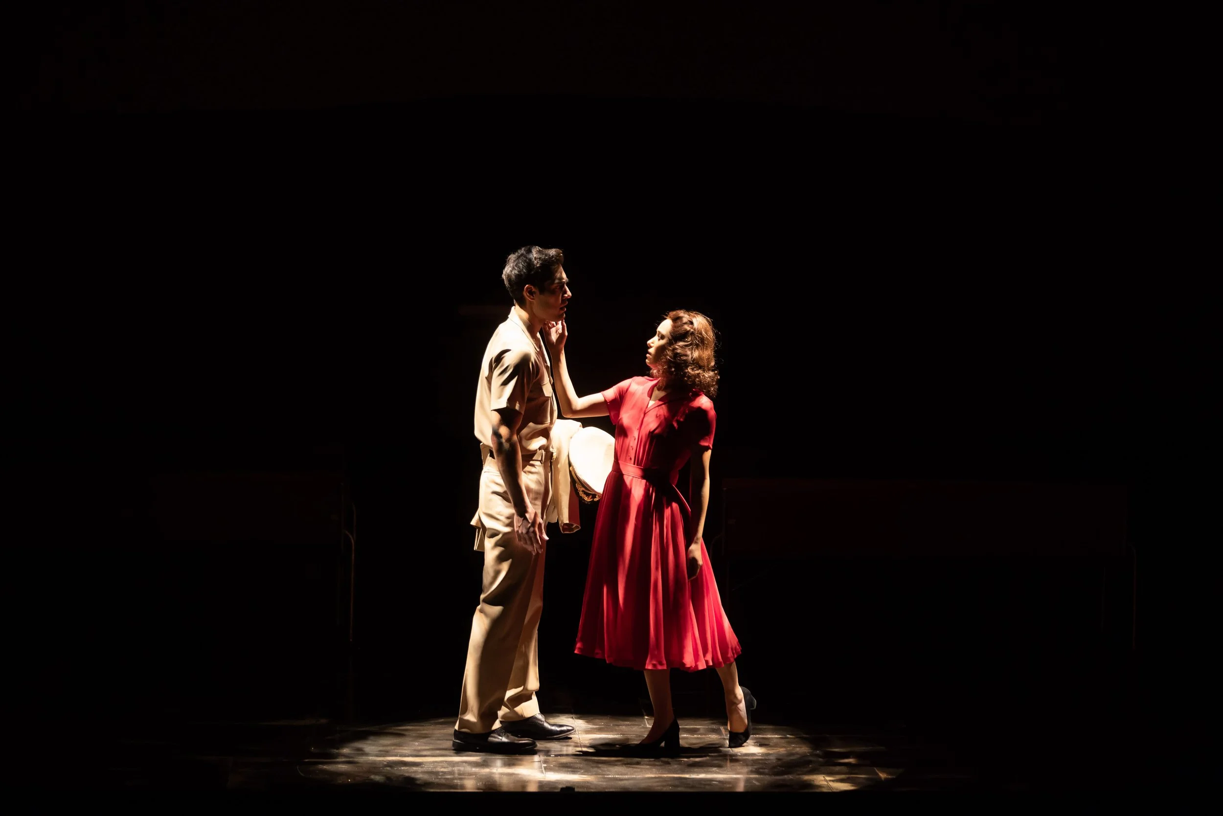 Two actors, a man and a woman, are on a dark stage under a spotlight. The man is wearing light-colored clothing and glasses, while the woman is dressed in a red dress with curly hair. They are engaging in an emotional scene with the woman touching th