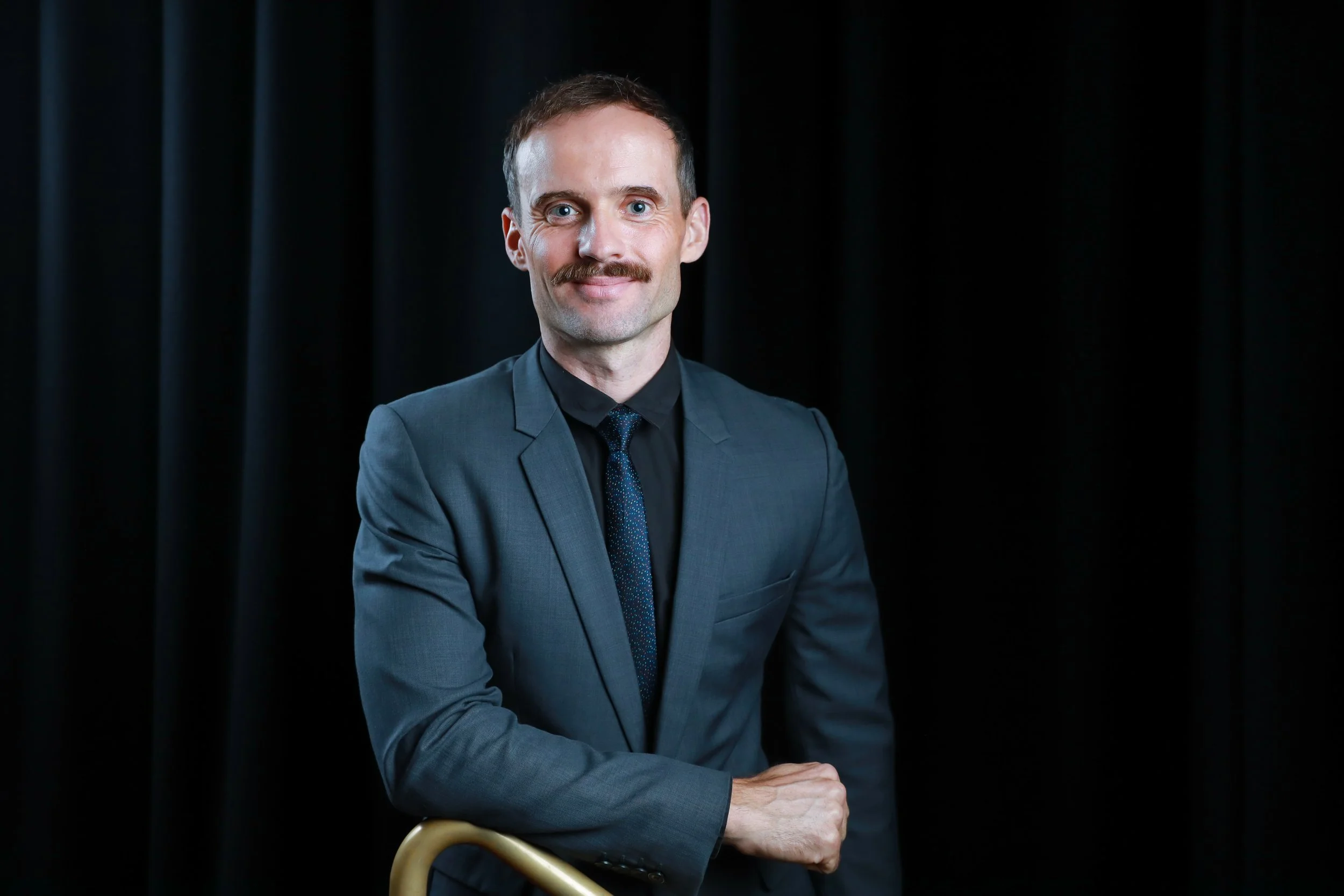 A man with light hair and a mustache wearing a gray suit, black shirt, and tie, posing with a smile against a black curtain background.