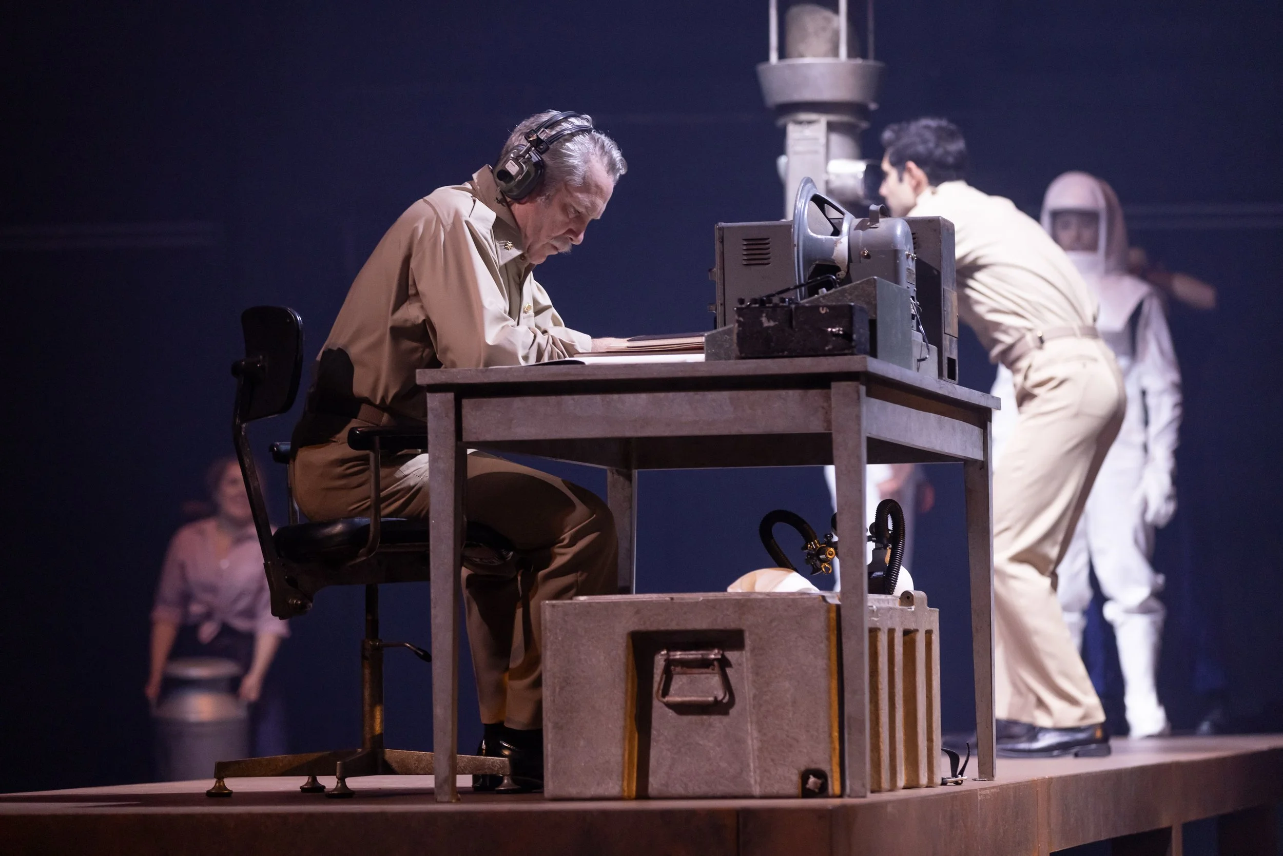 A man with headphones sowing at a desk with a large machine, with a woman in costume in the background.