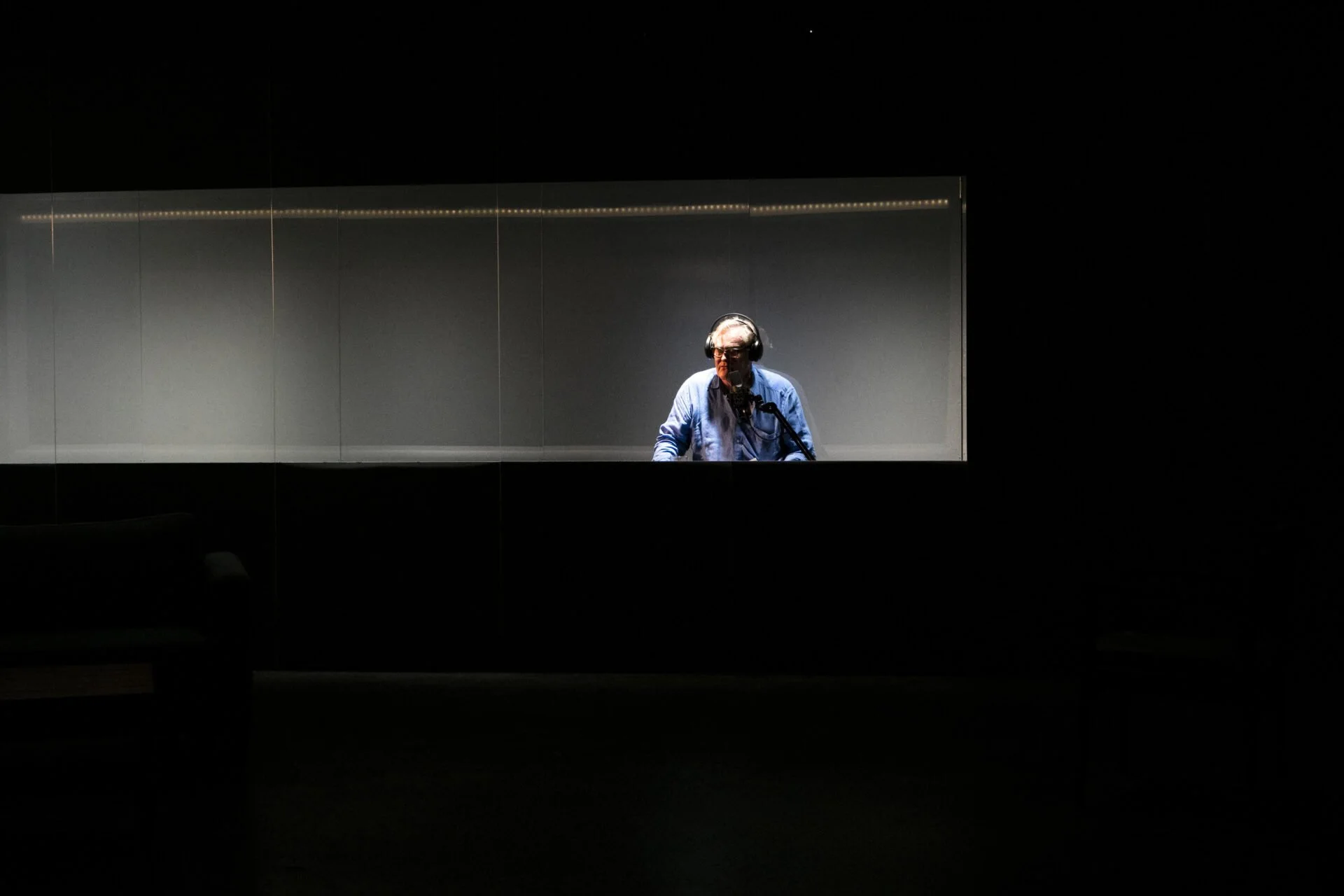 A man wearing headphones and glasses stands in front of a microphone inside a soundproof recording booth with gray walls and a long strip of lights on the ceiling.