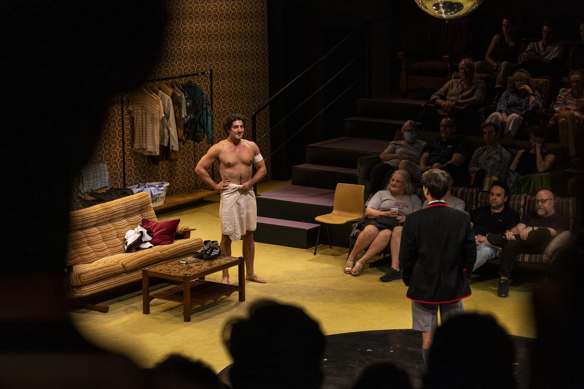 A shirtless performer on stage wearing a towel around his waist, smiling at the audience, with clothing hanging on a rack behind him. The audience, seated in tiered seating, watches him, with some wearing masks.