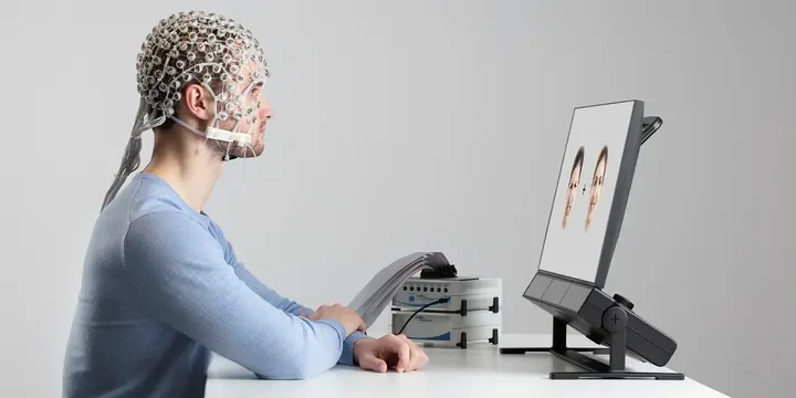 Person wearing a neuroimaging cap looking at a computer screen displaying images of eyes and eyebrows.